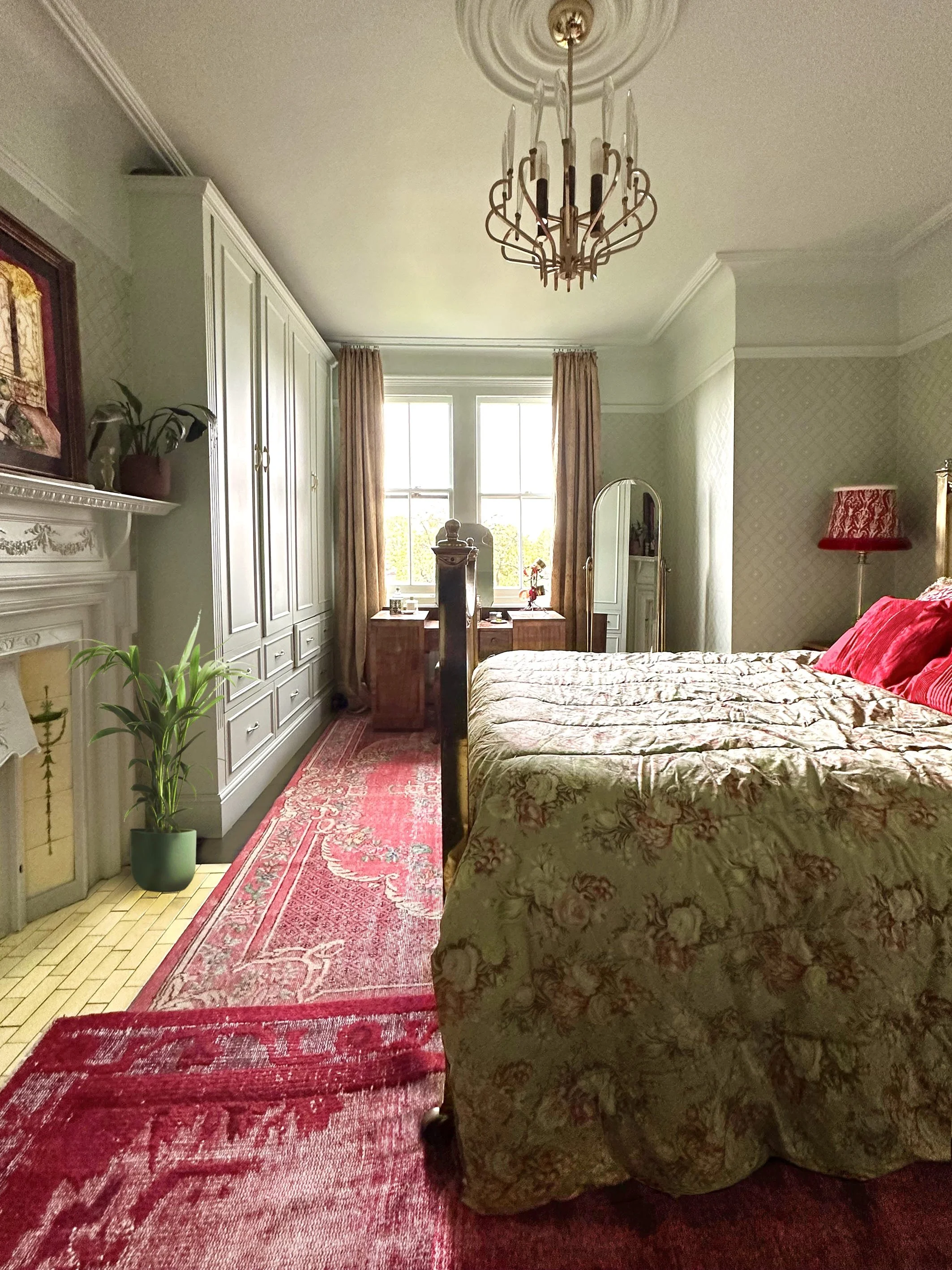 A bedroom with a large bed covered in a floral comforter and red pillows, a chandelier hanging from the ceiling, a window with curtains, a mirror, a wooden desk, a potted plant, and a red rug on a parquet floor.