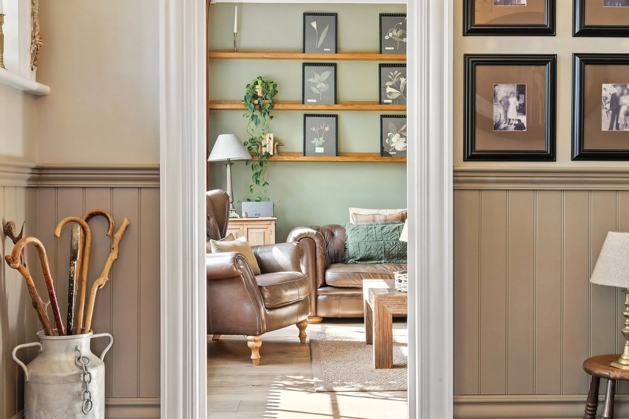 View of a cozy living room through an open doorway, featuring brown leather armchairs, a wooden coffee table, a green throw blanket, a floor lamp, and framed botanical art on the walls.