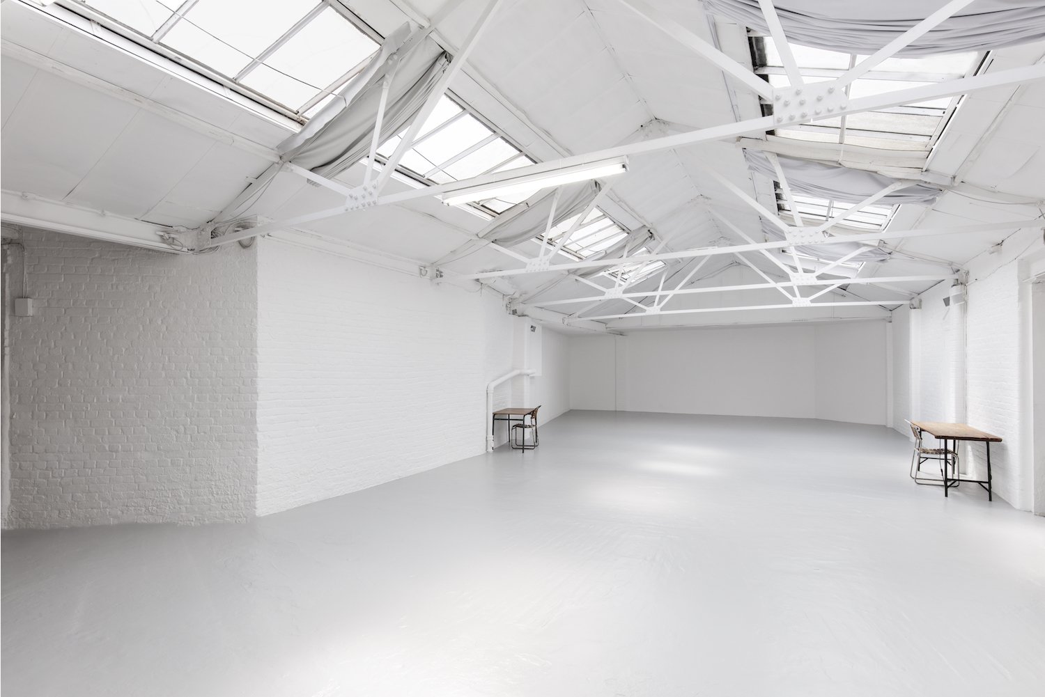 An empty, whitewashed industrial-style room with a high ceiling featuring exposed beams and skylights. There are two small tables with chairs, one on each side of the room.