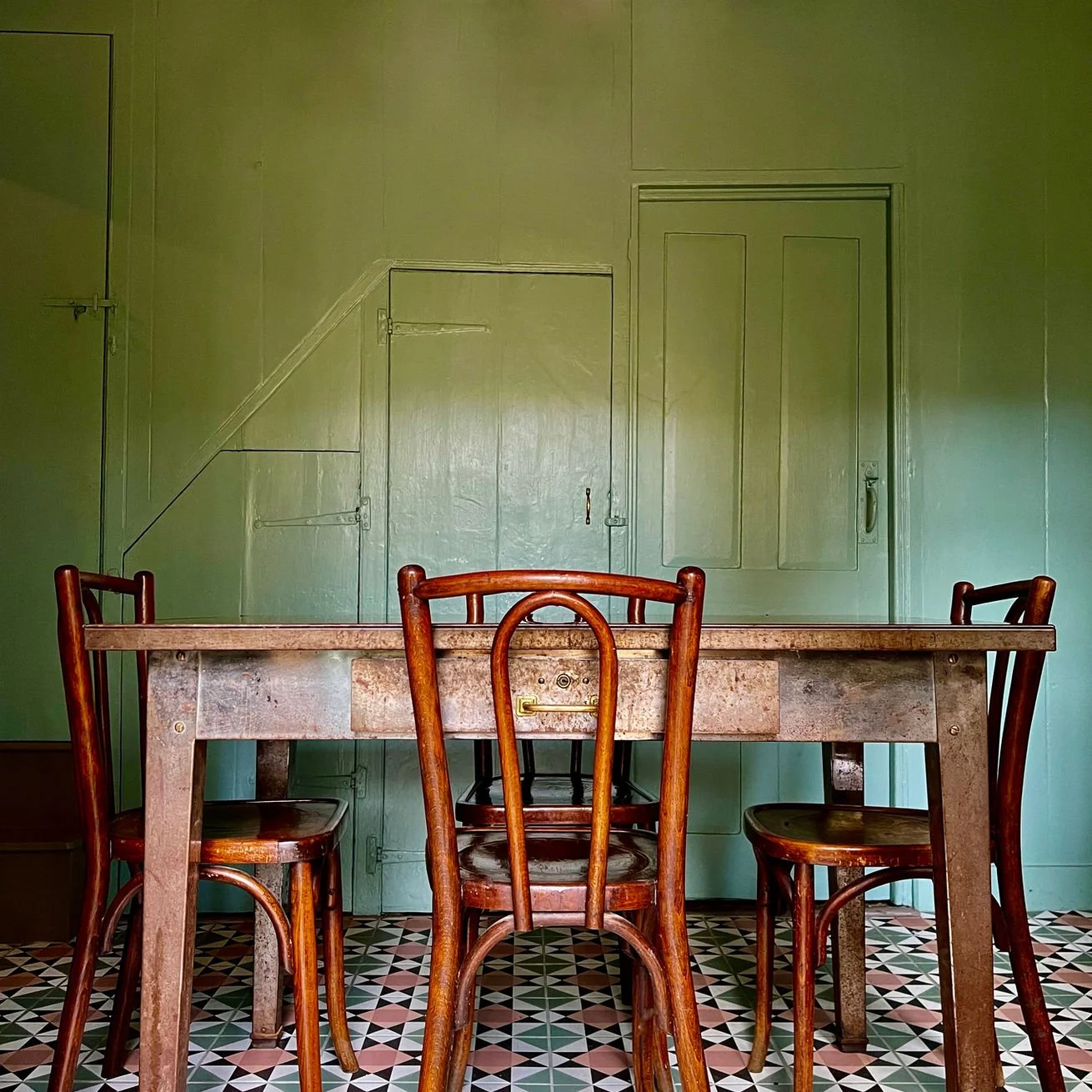 A rustic dining room with a wooden table and four wooden chairs, set against a green wall with two doors and an angled panel.
