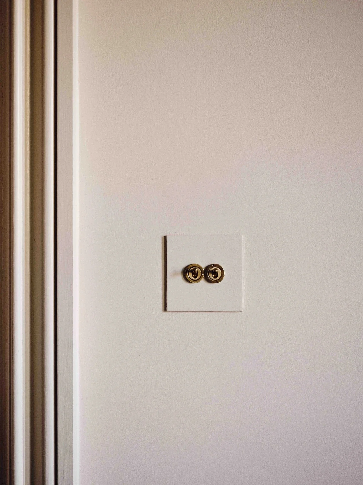 Close-up of a white electrical outlet cover with two brass wall switch toggles on a beige wall, with part of a wooden door frame on the left.