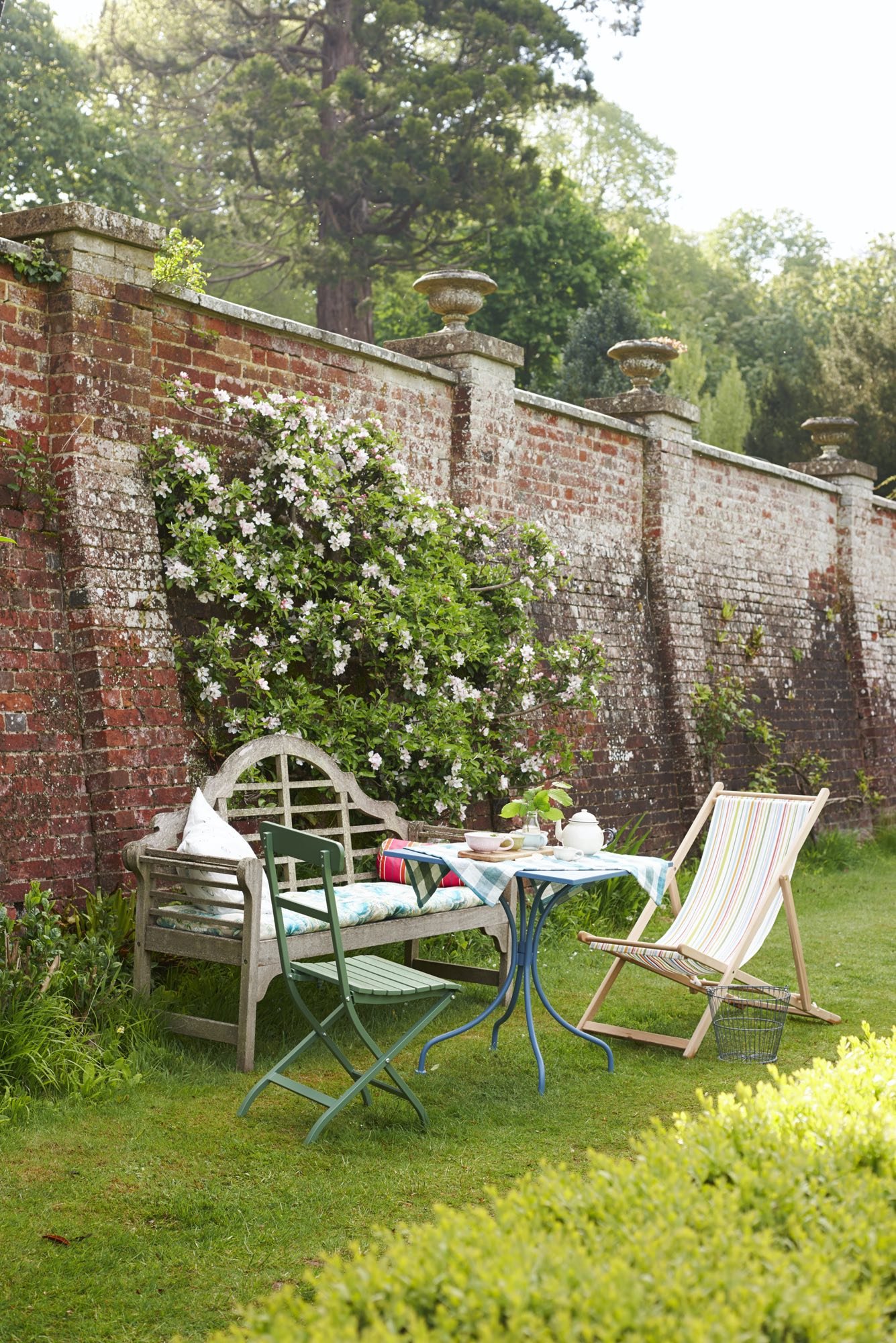 A cozy outdoor patio setup with a wooden bench, a green metal chair, and a striped beach chair arranged around a small round table with a checkered tablecloth, tea cups, a teapot, and a small plant, all set against a weathered brick wall with plants 