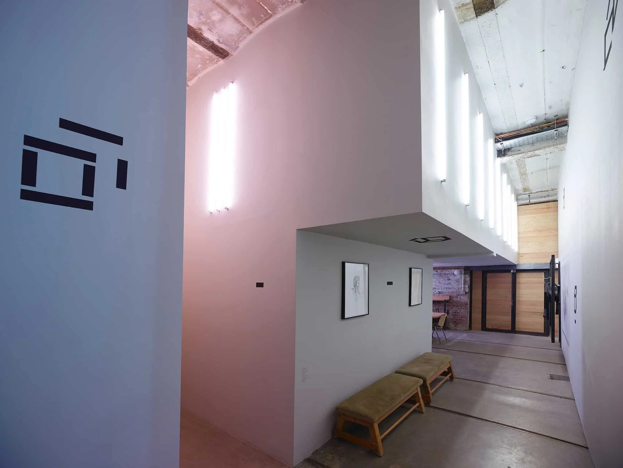 Interior of an art gallery with white walls, modern lighting, framed artwork, wooden benches, and a glass door at the back.