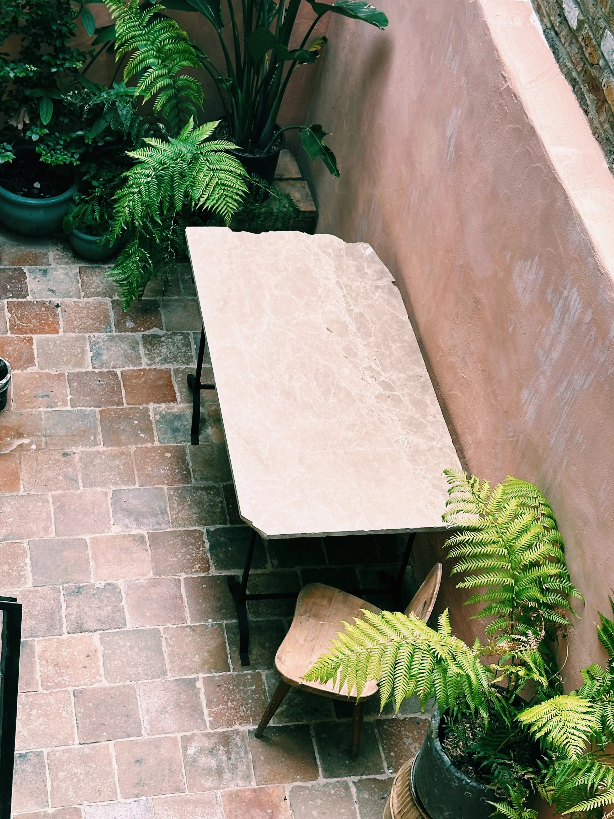 A small seating area with a marble-topped table, a wooden chair, and several potted green plants on a brick floor next to a pink stucco wall.