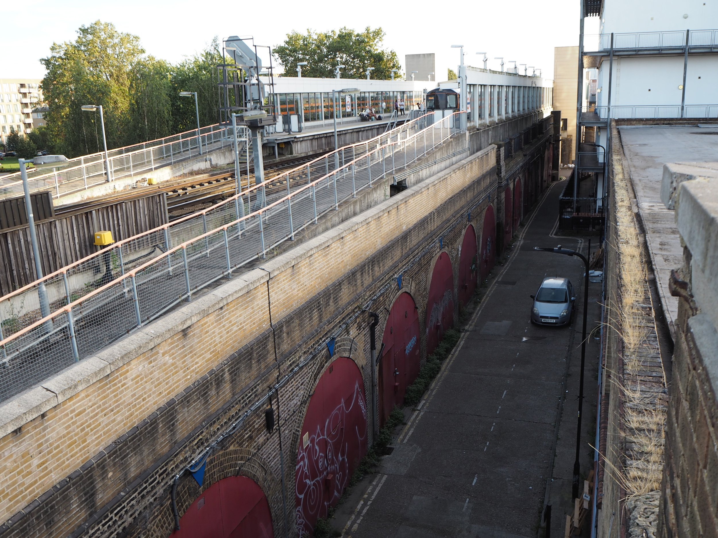 An urban scene showing a tram station with a train on the tracks, an escalator, and a parking area below with a car parked. The road is lined with streetlights and the station is surrounded by trees and multi-story buildings.