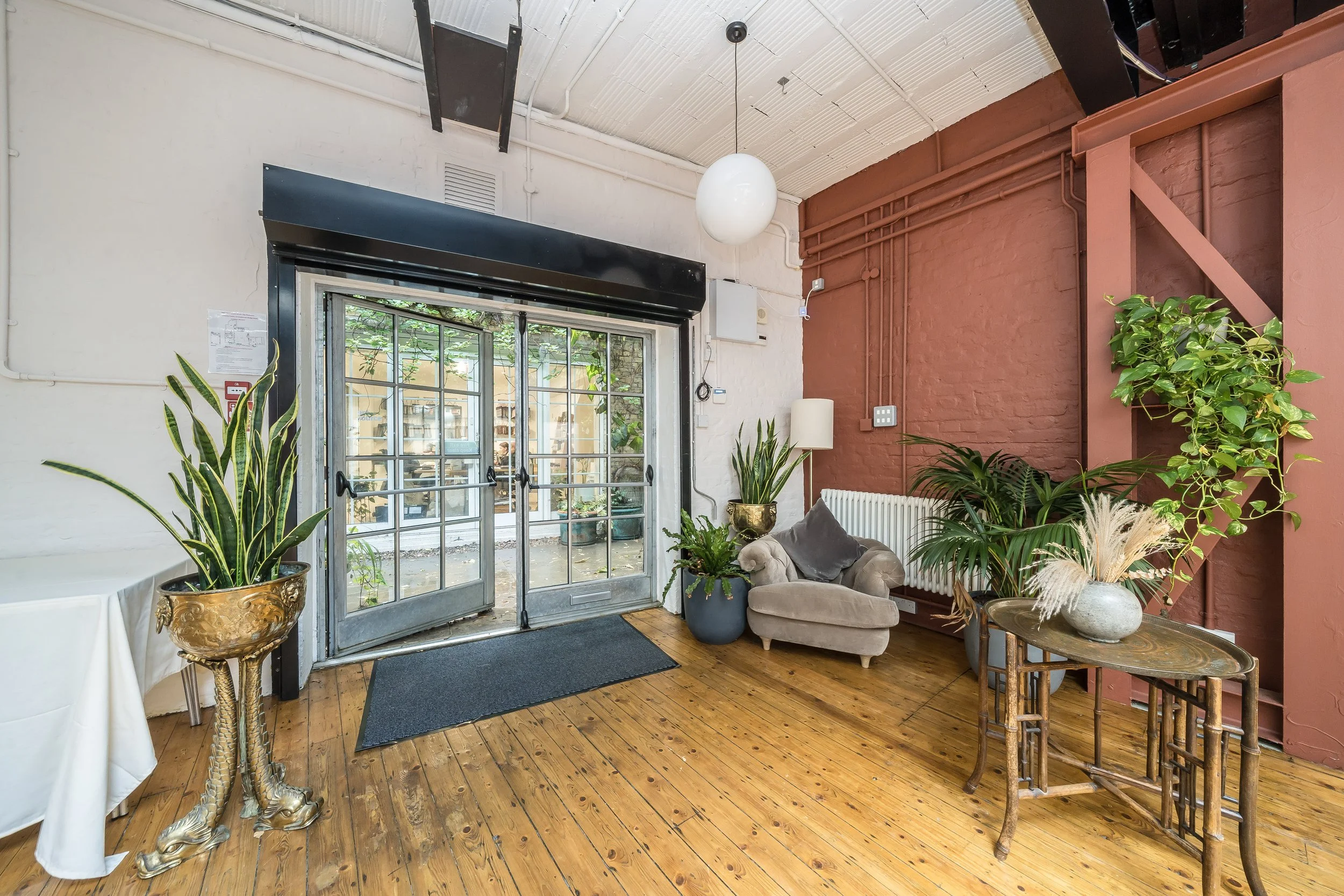 Cozy indoor space with wooden flooring, decorated with lush green plants in decorative pots, a beige armchair, a white radiator, a brown wooden table, and vibrant painted walls, with a glass door leading outside.