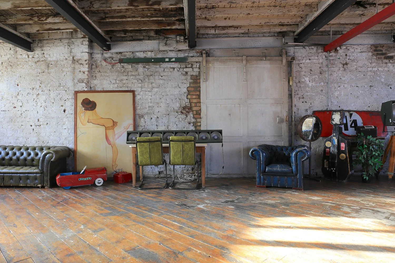 Interior of an industrial-style room with wooden floors and exposed brick walls. There are two vintage leather chairs, a poster of a woman, a small play car, a desk with two green chairs, and retro arcade games.
