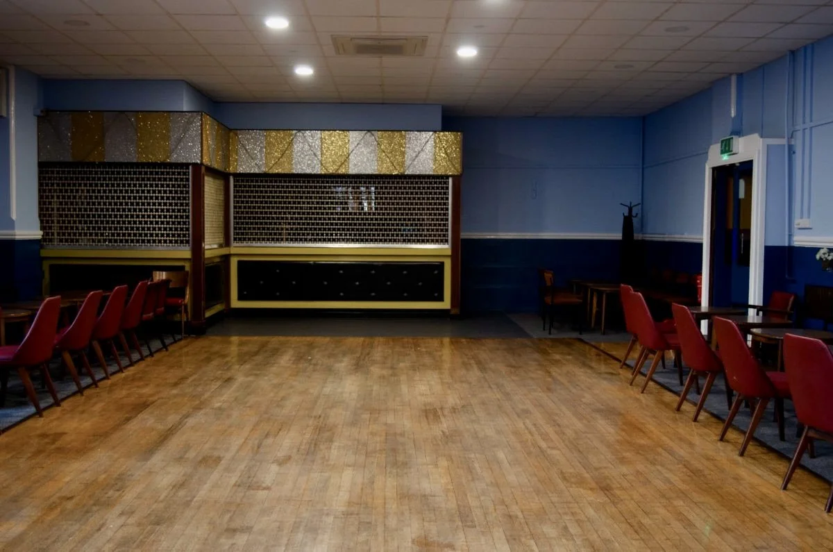 Empty event space or banquet hall with a wooden dance floor, red chairs lined up along the sides, decorative gold and silver paneling on the back wall, and blue walls with a coat rack near a door.