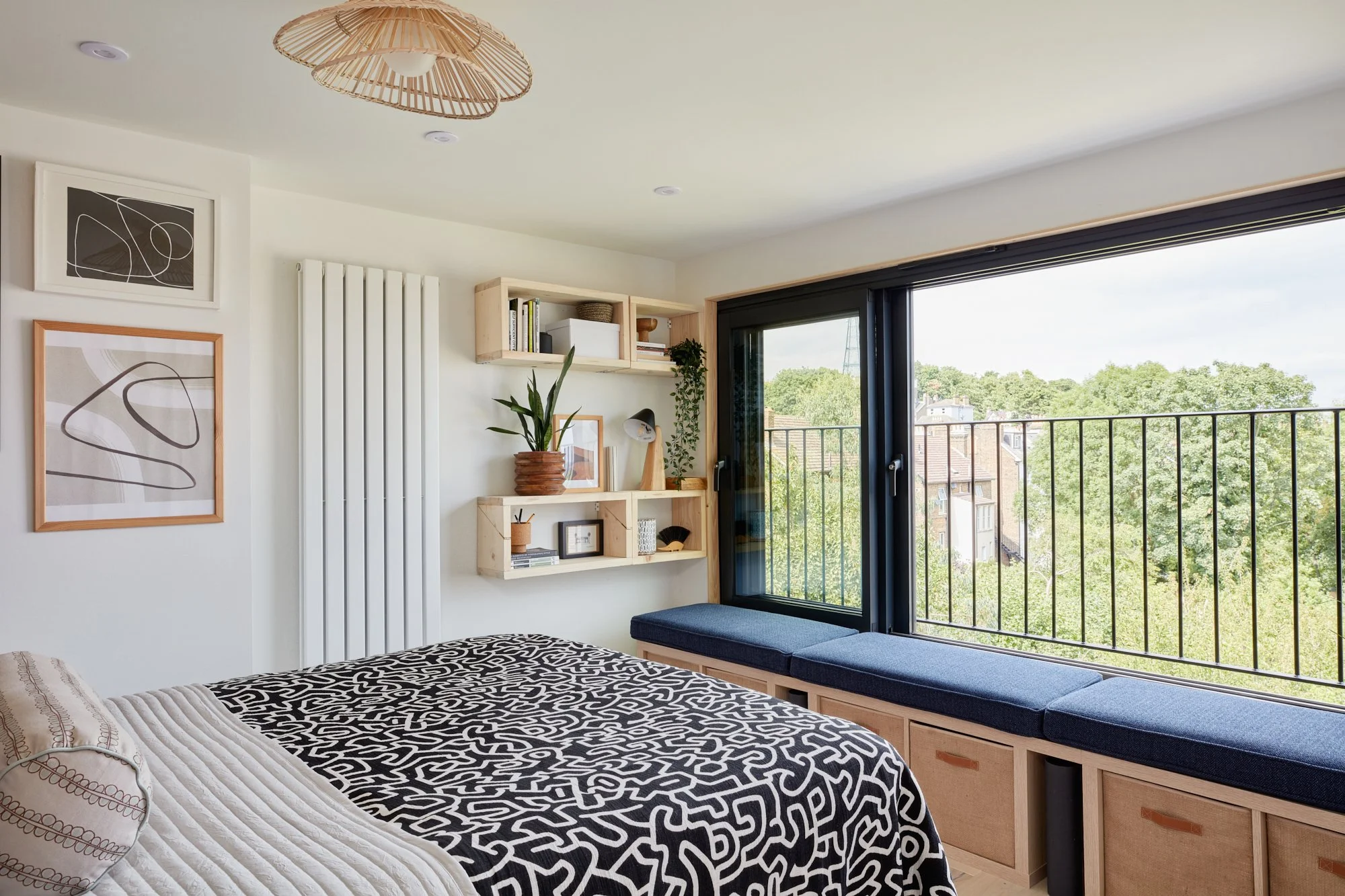 Bedroom with a large window, black and white patterned bedding, wall art, wooden shelves with decor, and a window seat with blue cushions.