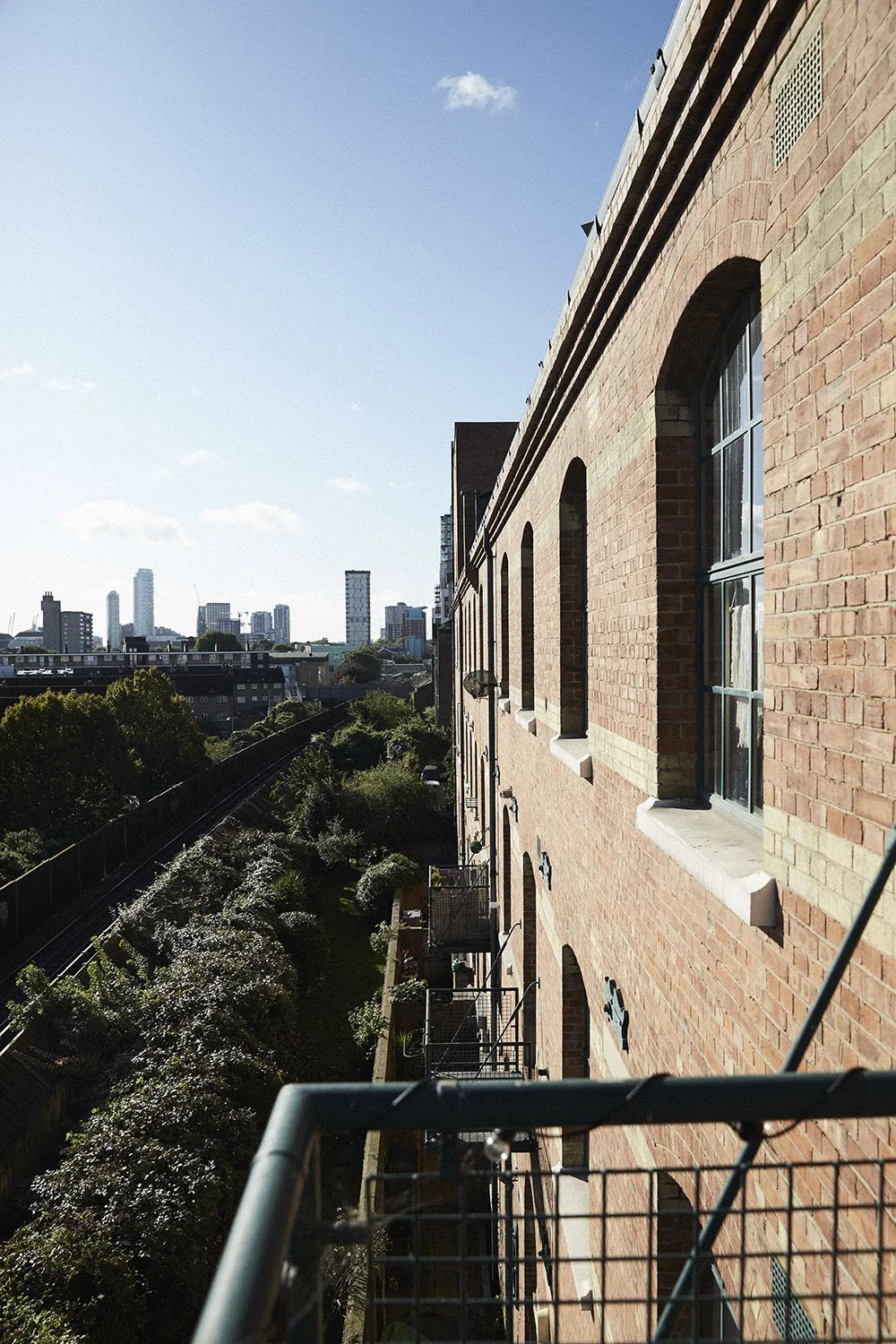 View of a brick apartment building with multiple windows and small balconies, looking down a city street with trees and tall buildings in the distance under a blue sky.