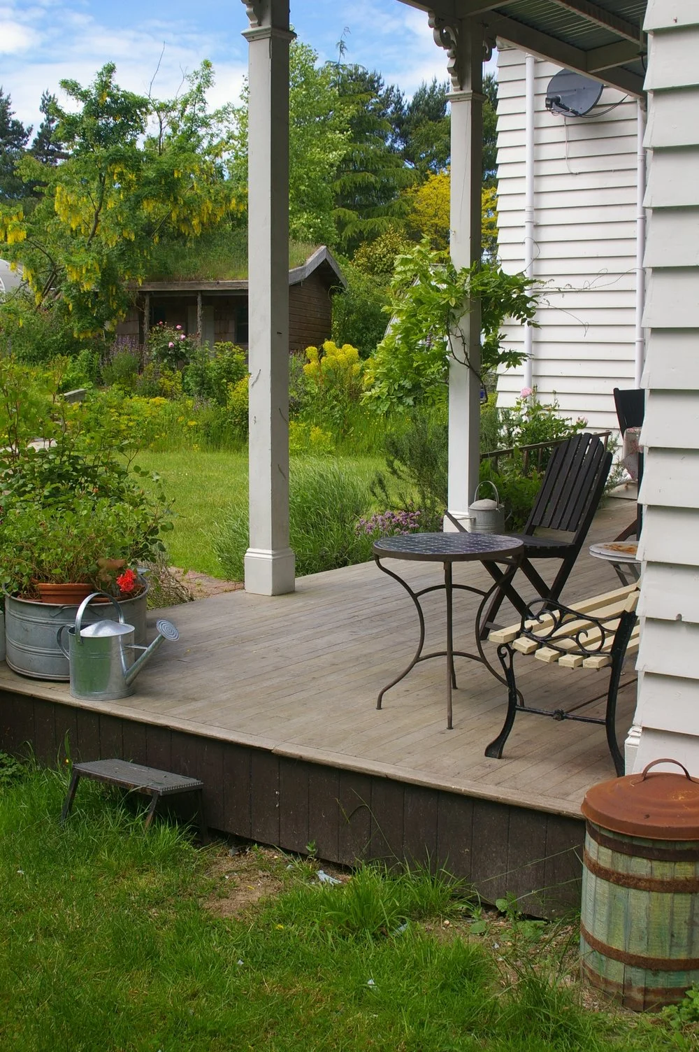 A cozy porch with black outdoor chairs and a small round table, surrounded by lush greenery and flowering plants.