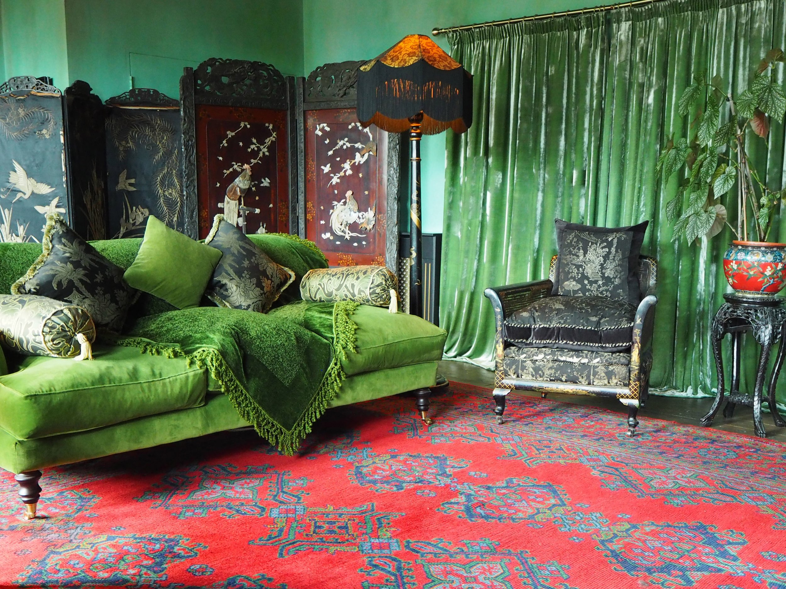 A vintage living room with a green velvet couch, decorative pillows, a black armchair, a tall floral plant, and a red patterned rug with green curtains and a decorative folding room divider with Asian-style art in the background. A floor lamp with a 
