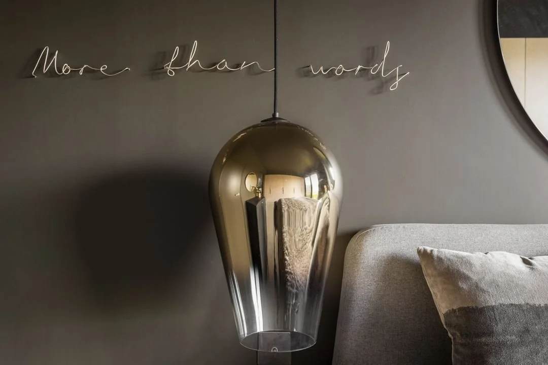 Close-up of a modern bedroom wall with a metallic pendant lamp, gray cushion, and a decorative wall sign that says 'More than words' in cursive.