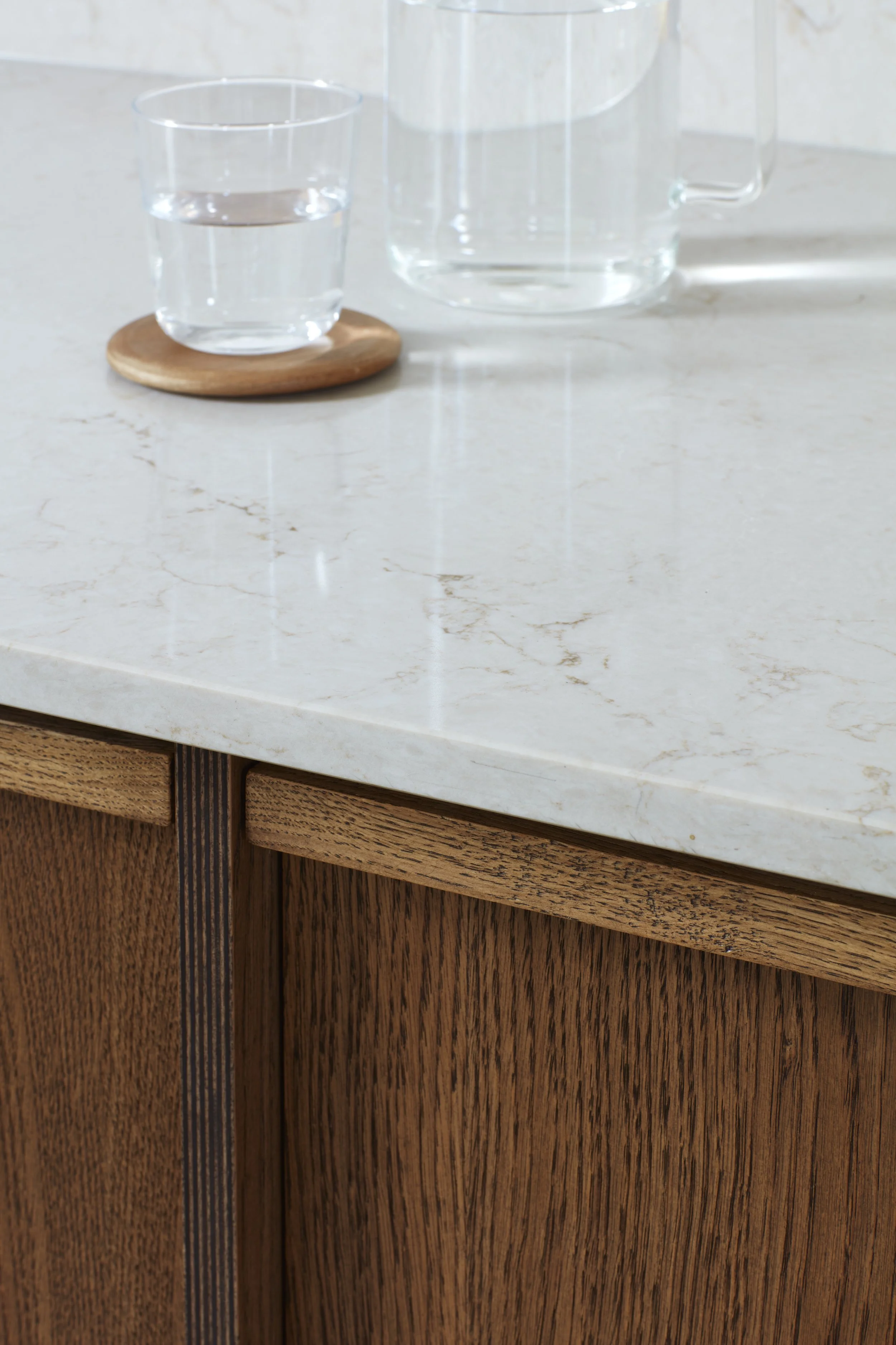Close-up of a light-colored marble countertop with wooden cabinet below, a glass of water on a round wooden coaster, and a glass pitcher in the background.