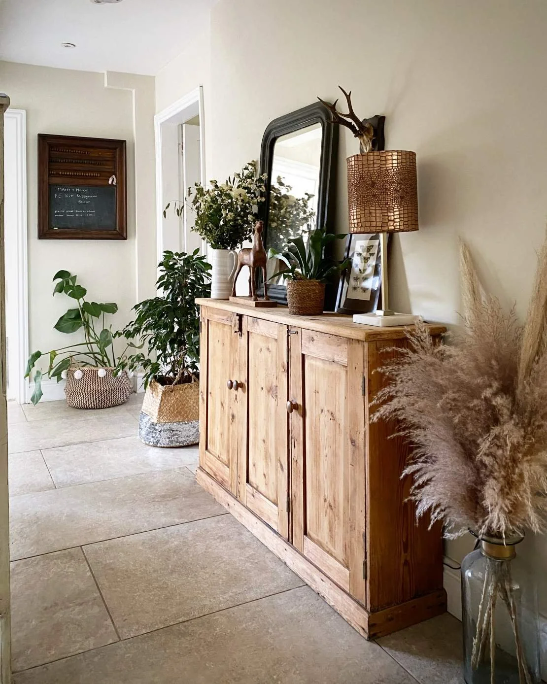 A wooden sideboard with plants and decorative objects, a tall glass vase with pampas grass, and a mirror reflecting part of the room. A table lamp with a woven shade is on the sideboard.
