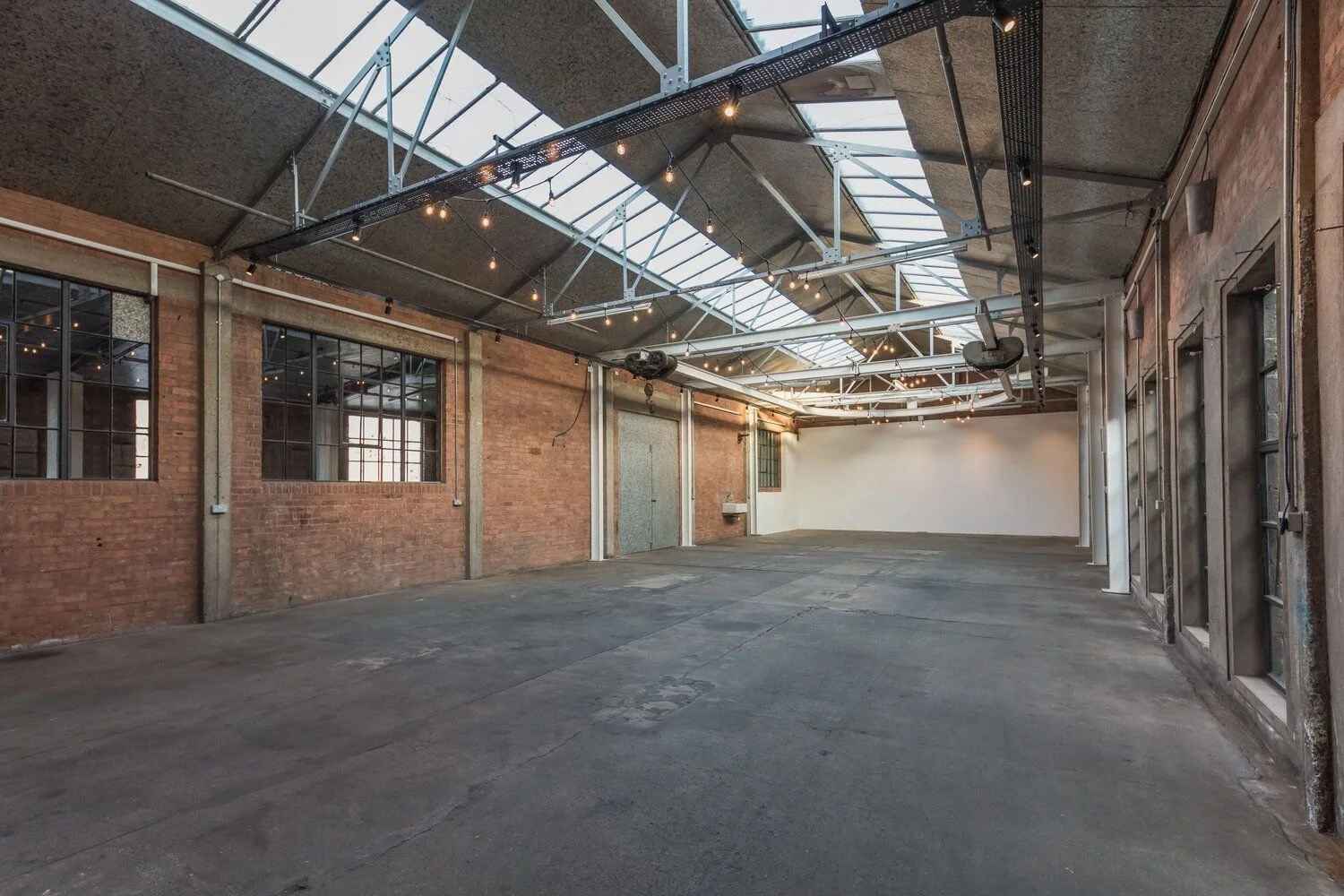 An empty industrial-style room with brick walls, large windows, and a concrete floor. The ceiling features a glass skylight, hanging string lights, and exposed metal beams.