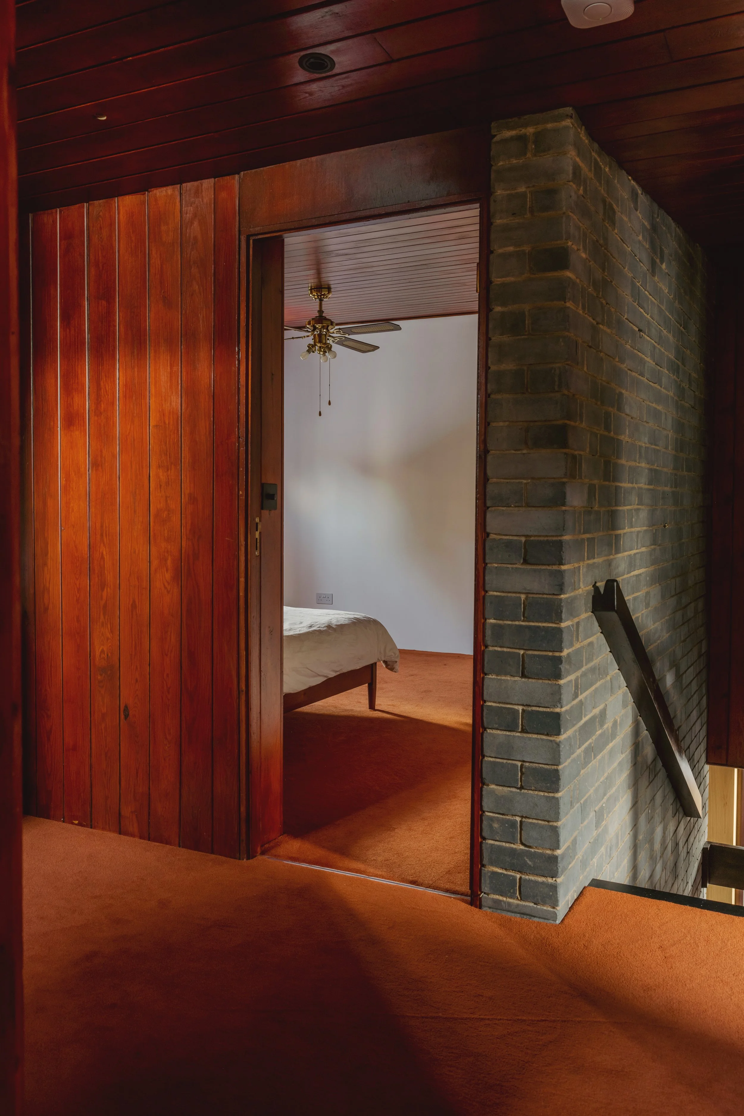 A doorway leading into a bedroom with wooden paneling on the ceiling and door frame, and brick walls. The room has a bed with a white duvet and a ceiling fan. The floors are covered with orange carpet.