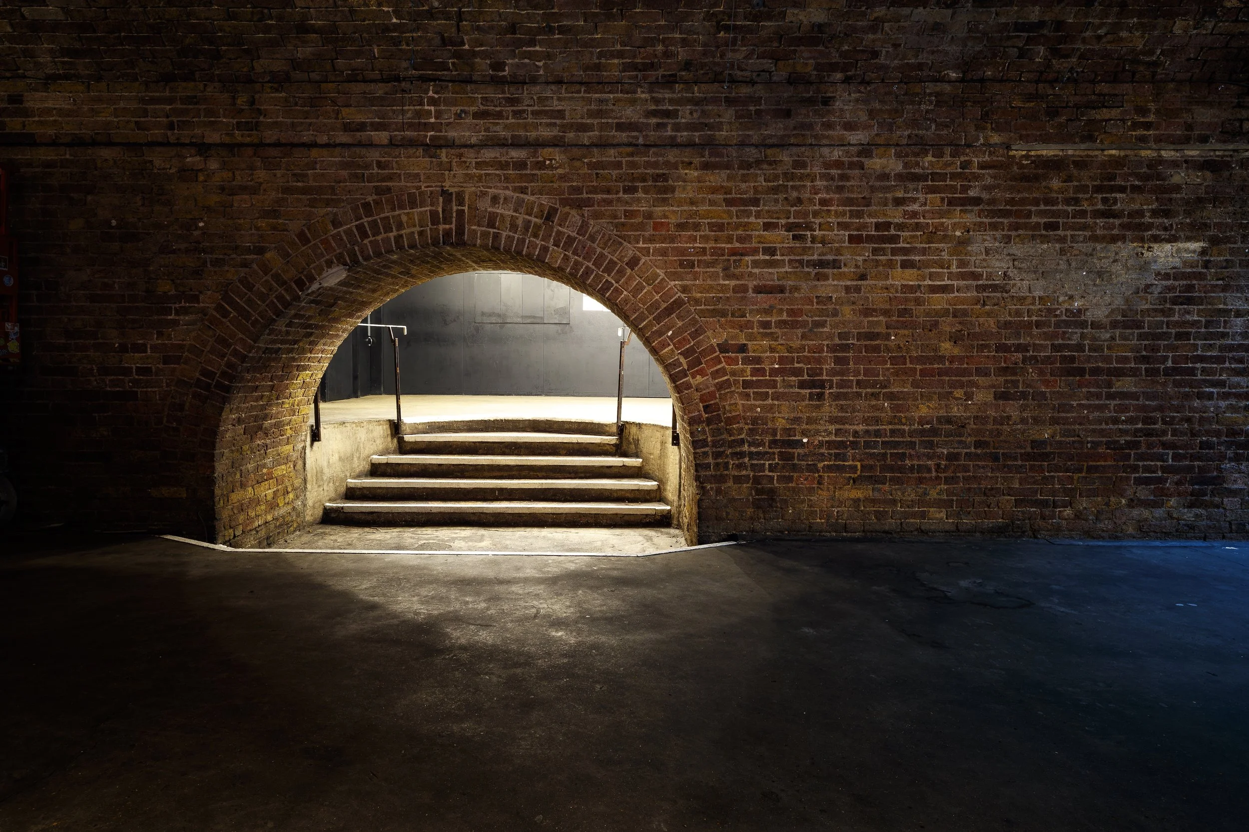 Brick wall with a small archway leading to a set of stairs, with handrails on either side, and a light illuminating the steps.