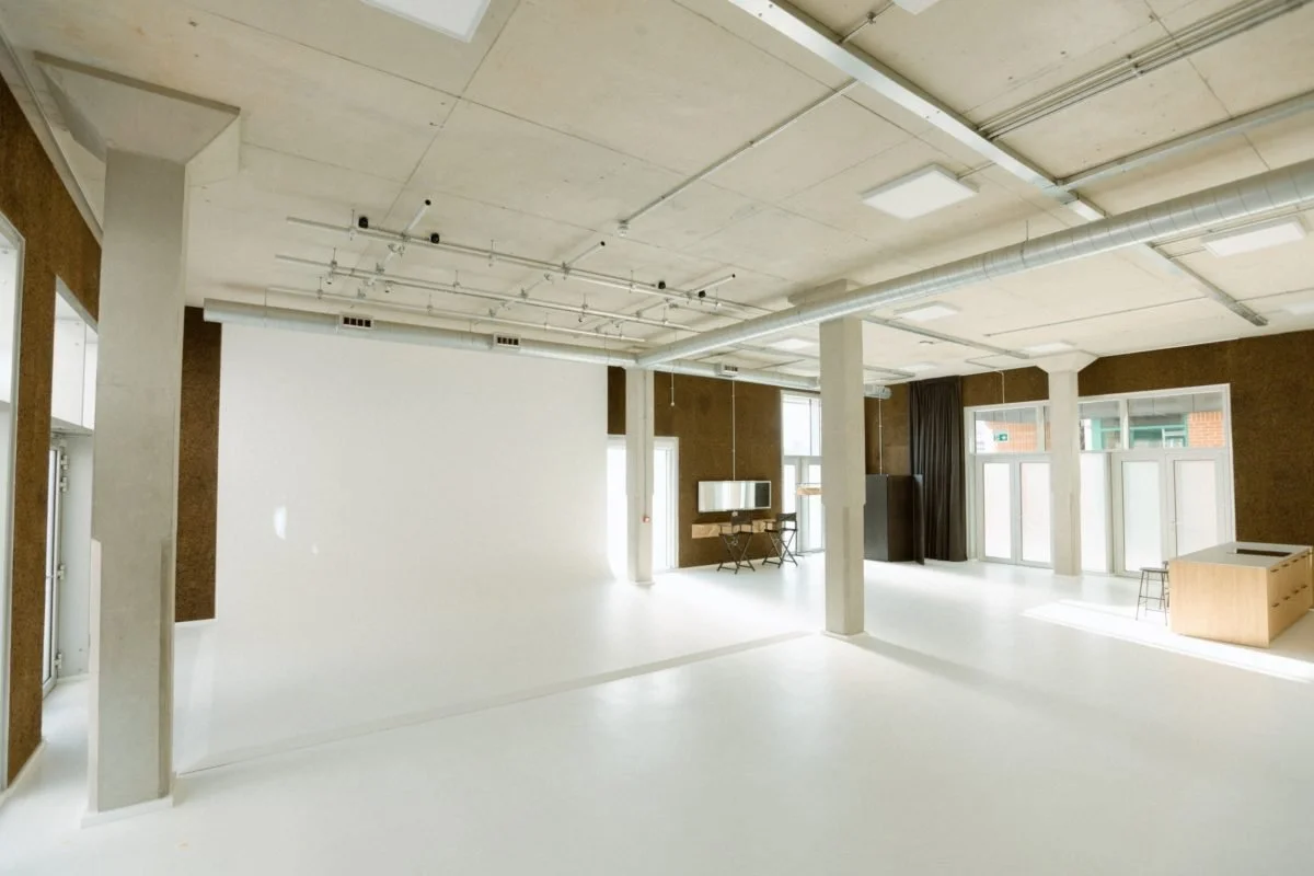 Empty modern room with large windows, white walls, exposed ceiling pipes, and minimal furniture.