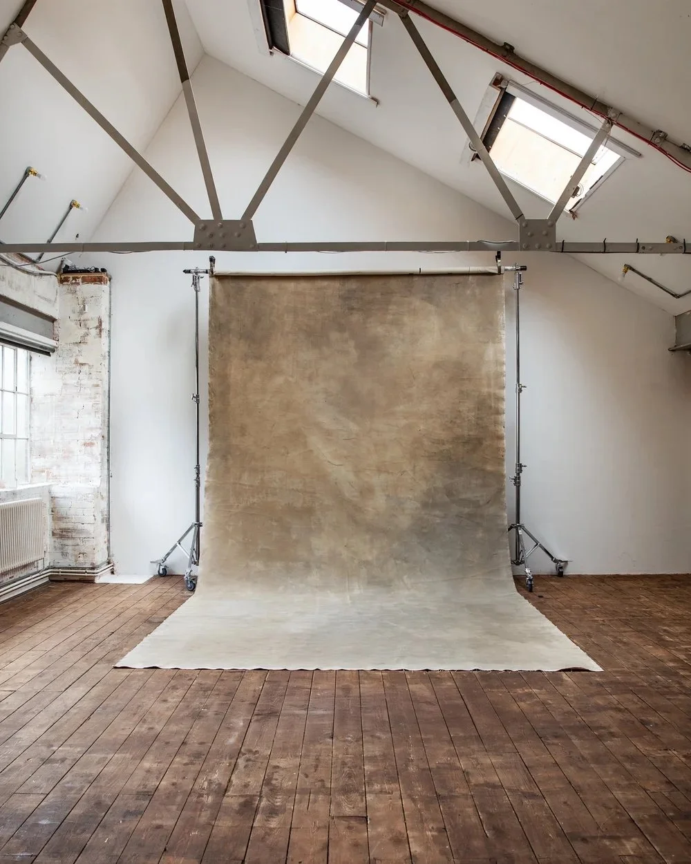 Indoor photo of a photography studio setup with a beige backdrop, wooden floor, large windows, and exposed brick wall.