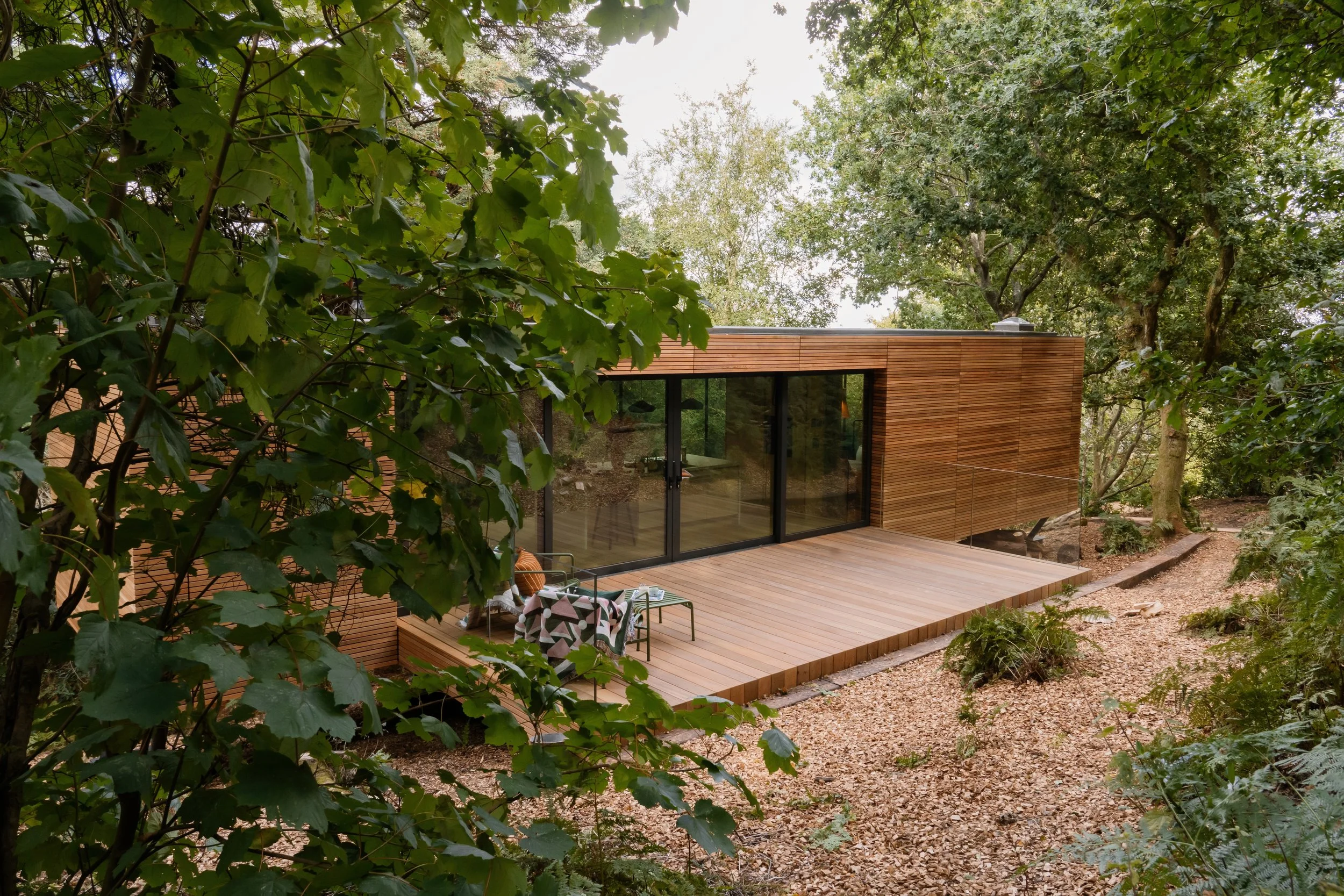 Modern house with wooden exterior and large glass sliding doors, surrounded by trees, with a wooden deck and outdoor seating area with chairs and a patterned blanket.