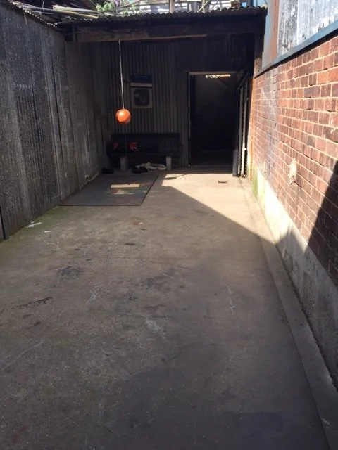 An outdoor alleyway or back yard with a concrete ground, fencing on the left, brick wall on the right, and a small covered area at the end with a hanging orange ball and a mat on the ground.