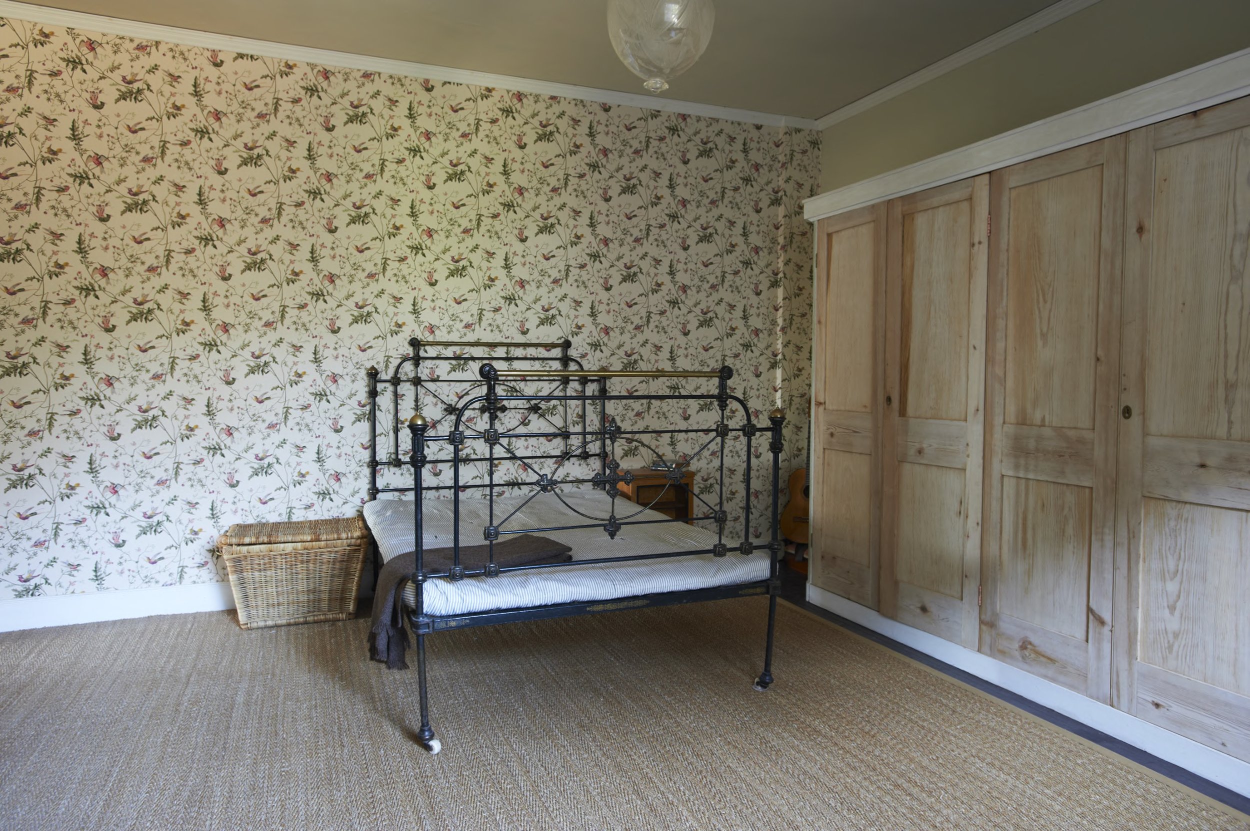 A bedroom with floral wallpaper, a black metal bed frame, a wicker basket, a wooden nightside table, a guitar, and a large wooden wardrobe.
