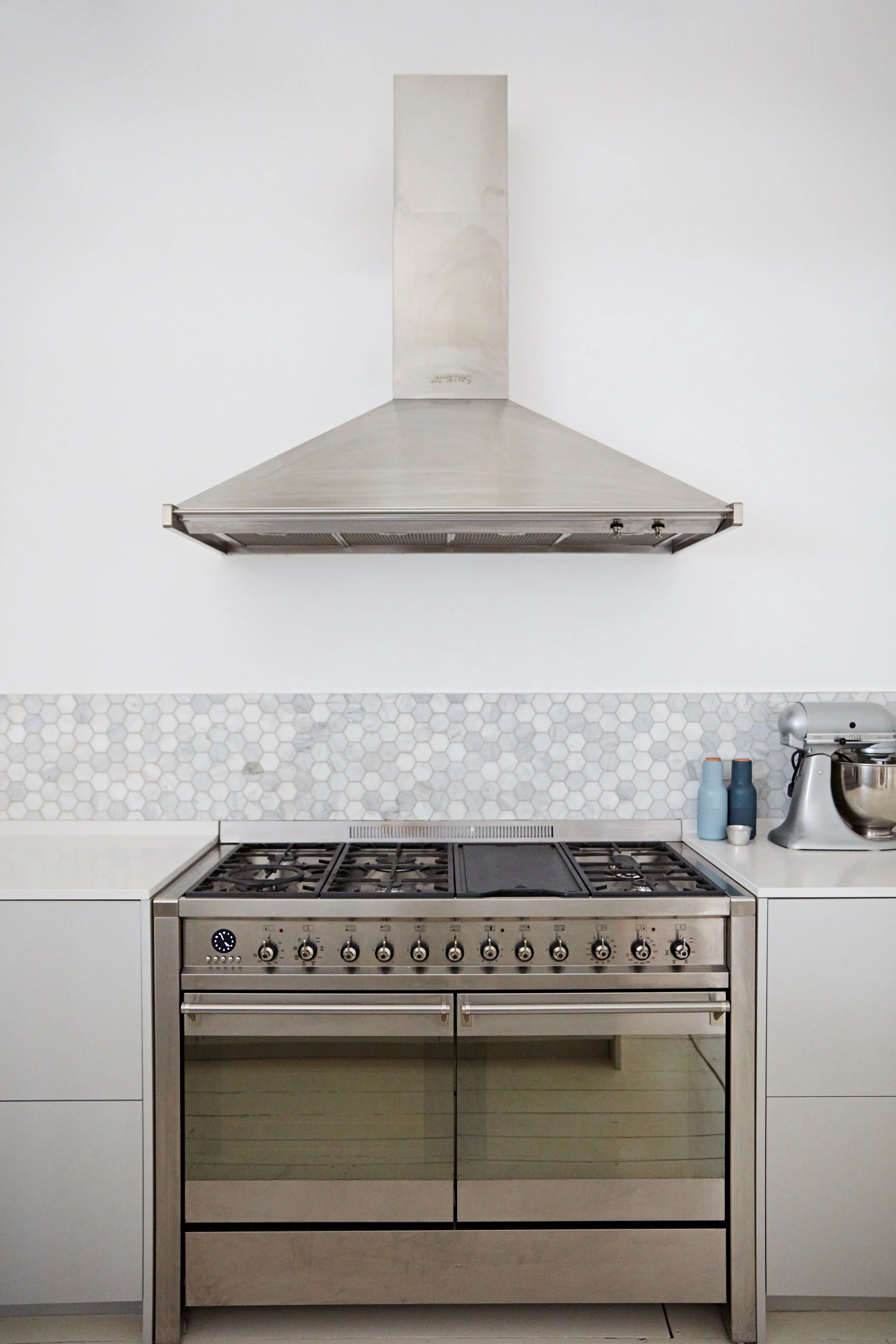 Modern kitchen with stainless steel stove and range hood, white cabinets, hexagon tile backsplash, and small appliances including a stand mixer and bottles.