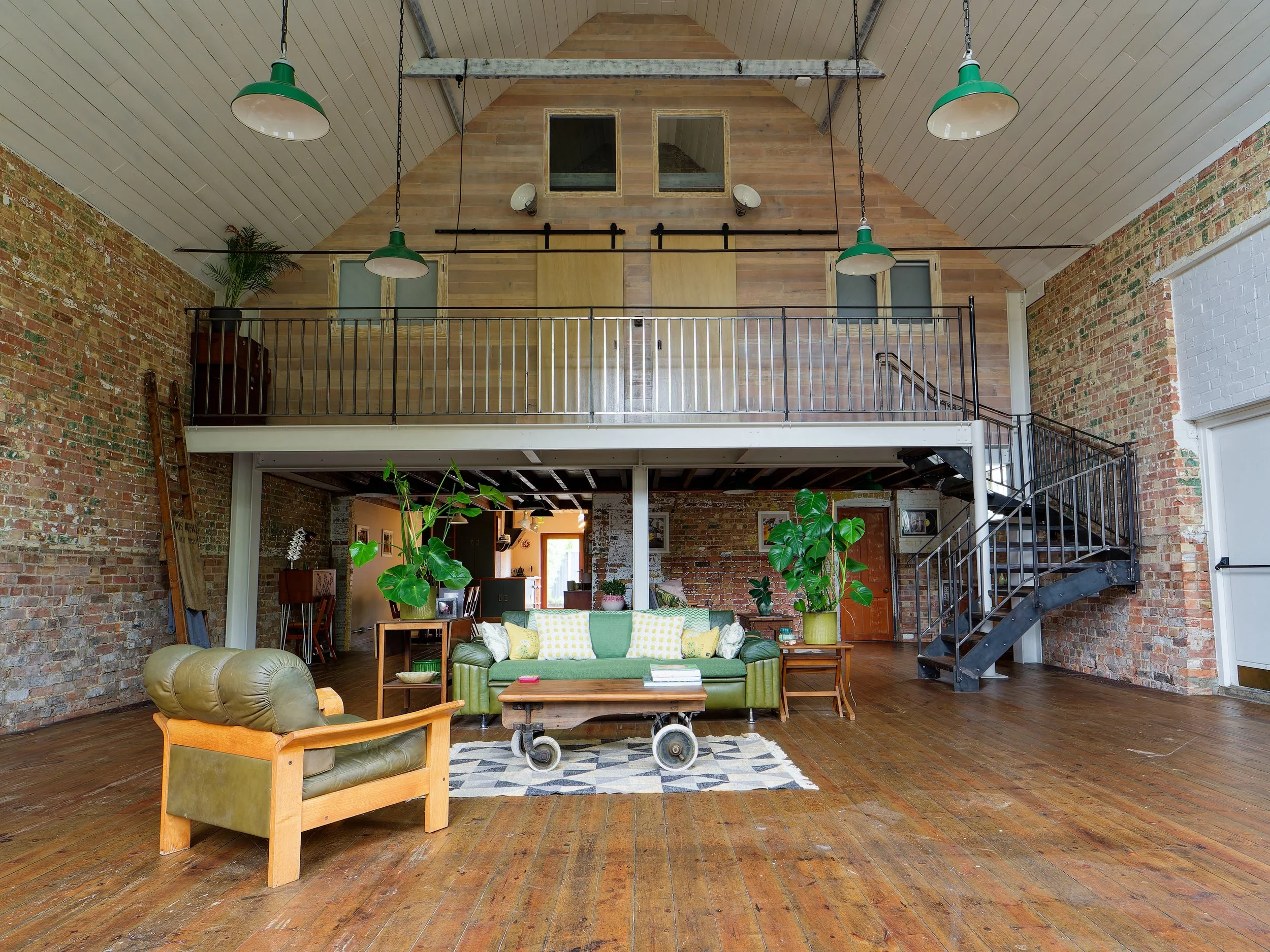 Open living room with exposed brick walls, wooden floors, and a loft area. There are green and beige sofas, large potted plants, and a wooden coffee table. Metal staircase leads to the upstairs loft, which has a wooden wall with windows and doors. Ov