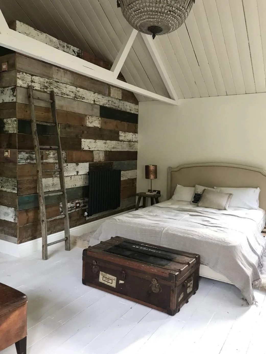 Rustic bedroom with a white bed, vintage trunk, wooden ladder, and a reclaimed wood accent wall.