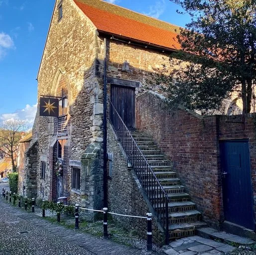 A historic stone building with a red-tile roof, an external staircase with metal railing leading to a door, a tree with green leaves on the right, and a street with sidewalk and bollards in the foreground, all under a clear blue sky.