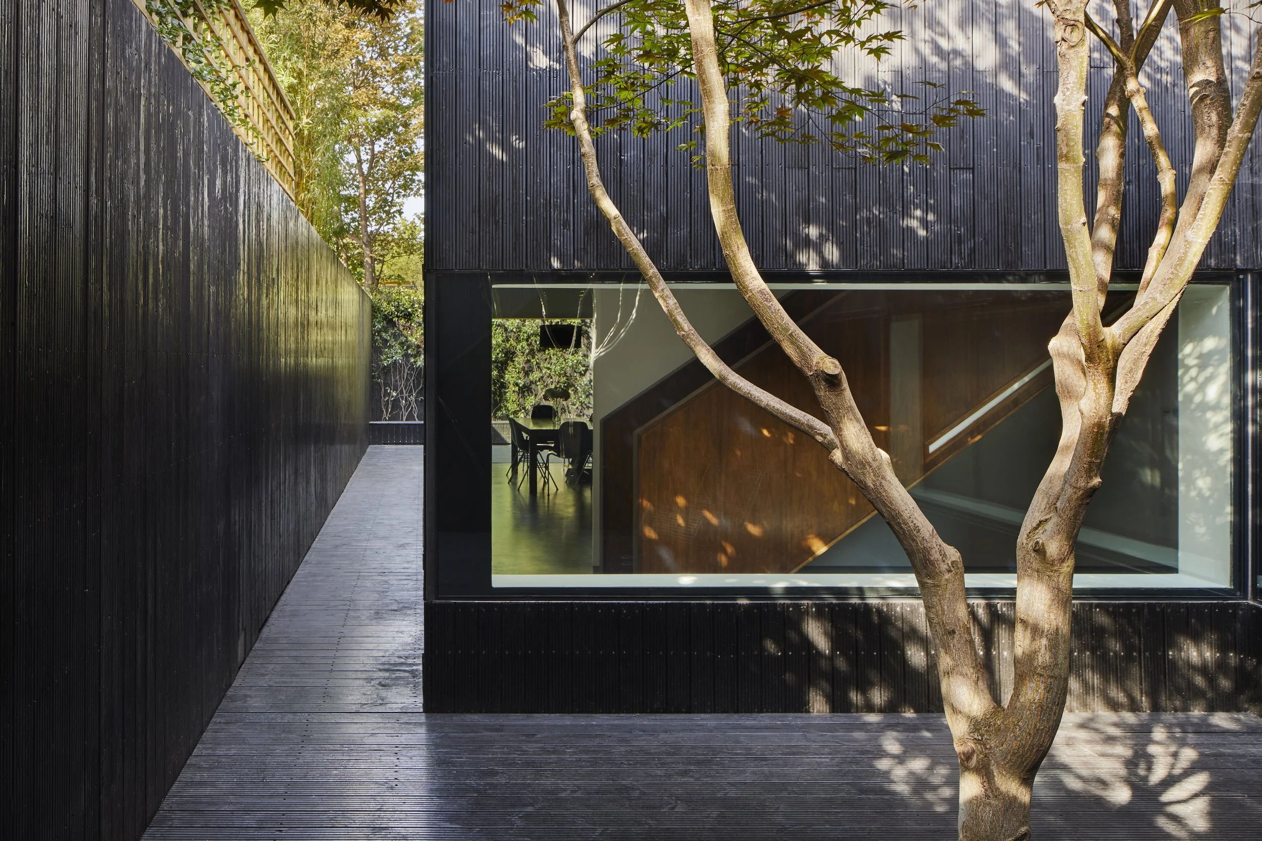 Modern building with black exterior walls, large window revealing a view of a dining area inside, and a tree in the foreground casting shadows on the wood deck.