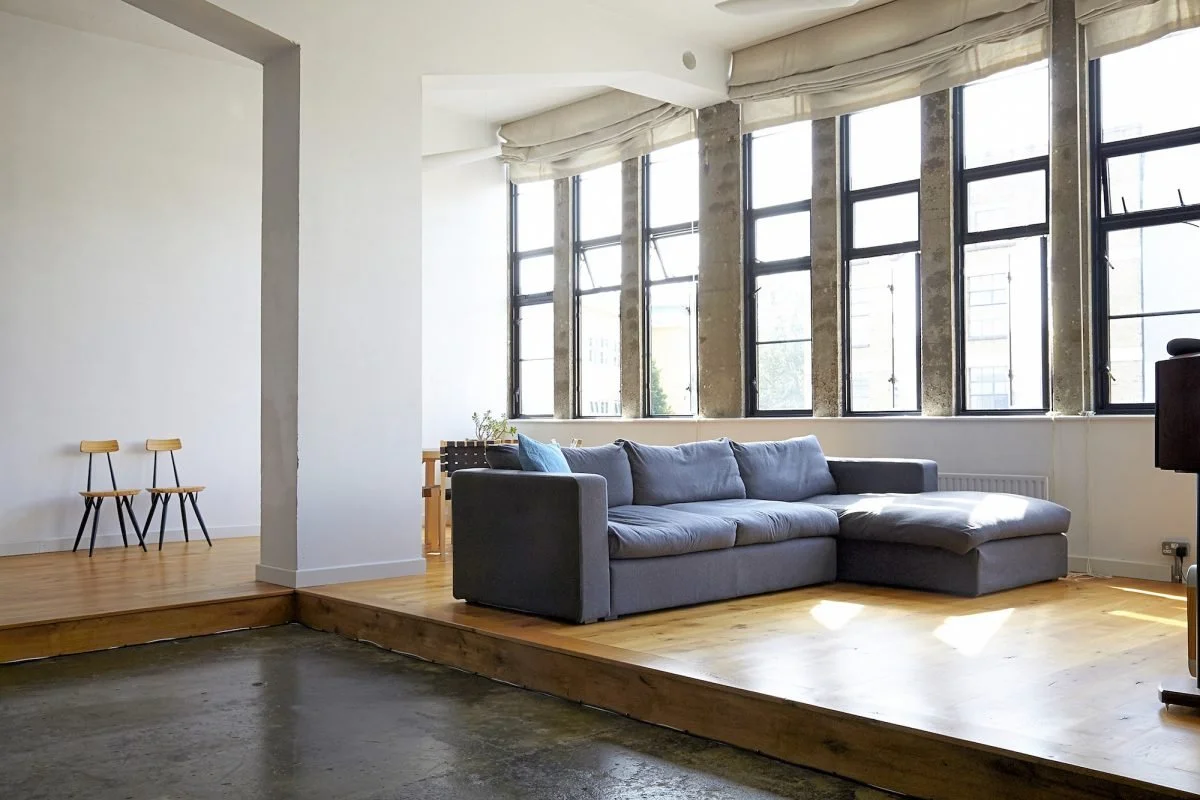 Modern living room with a gray sectional sofa, large windows with blinds, and wooden flooring.