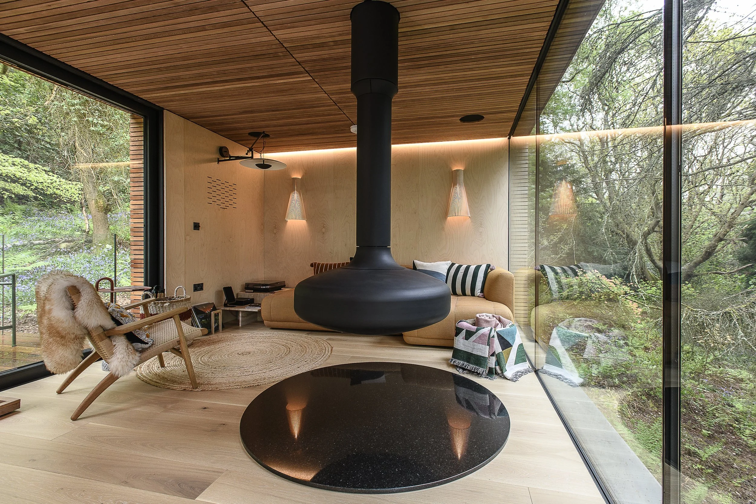 Modern living room with floor-to-ceiling glass walls, wooden ceiling and floor, a hanging fireplace, a beige sectional sofa with striped and patterned pillows, a wooden armchair with fur throw, a round black rug, and views of trees outside.