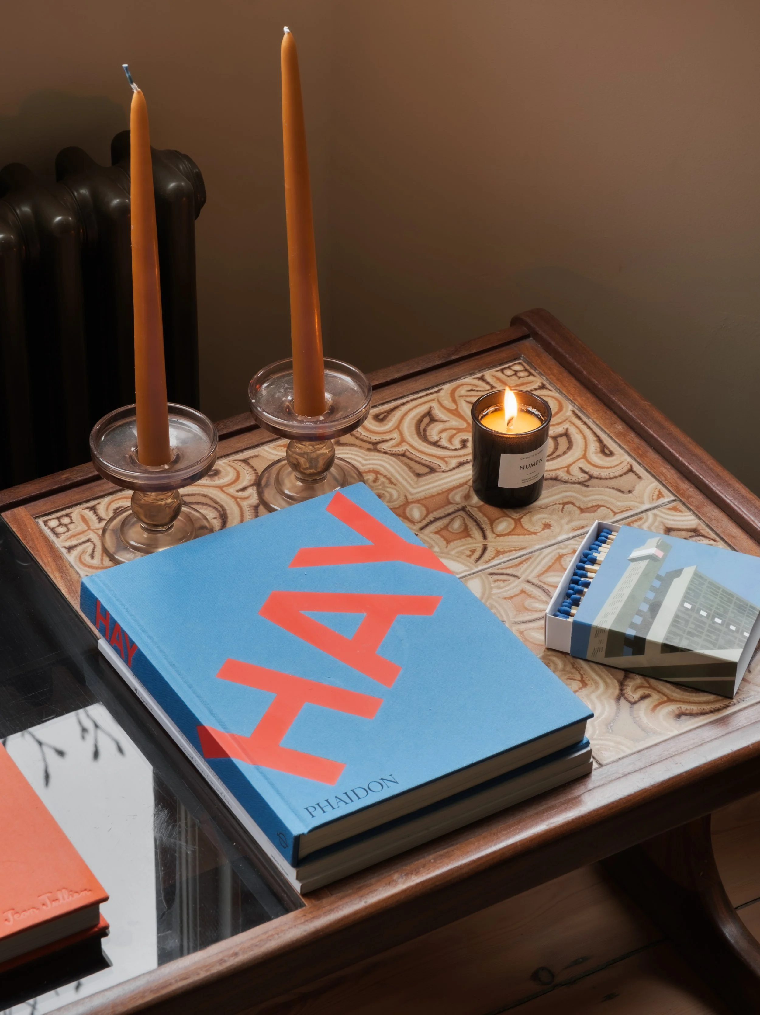 A wooden table with two tall, unlit candles in glass holders, a lit candle in a black container, a large blue book titled "HAY" by Phaidon, and a pack of blue and white matches.