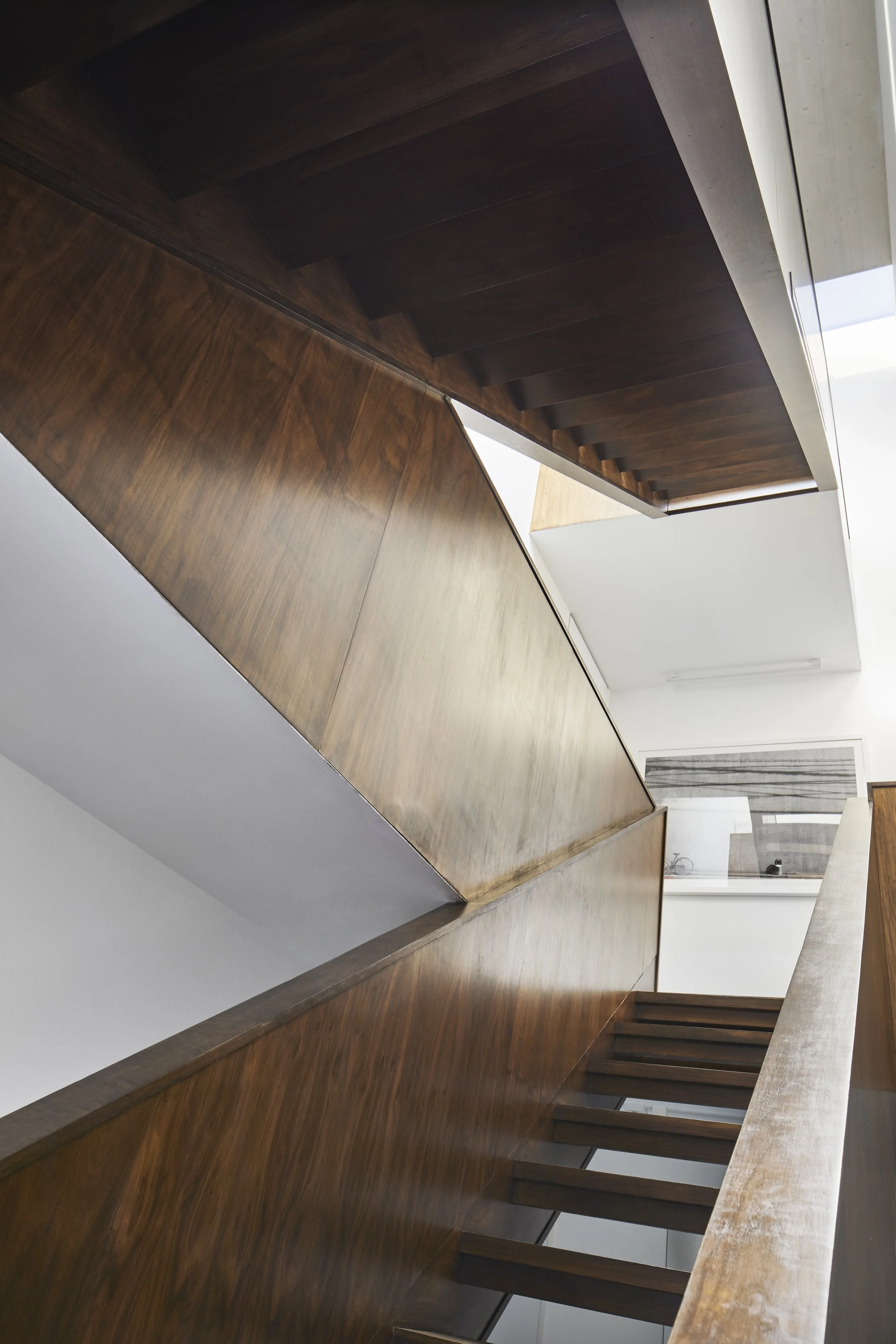 Interior view of a staircase with wooden steps and railings, viewed from below, with a window and modern decor in the background.