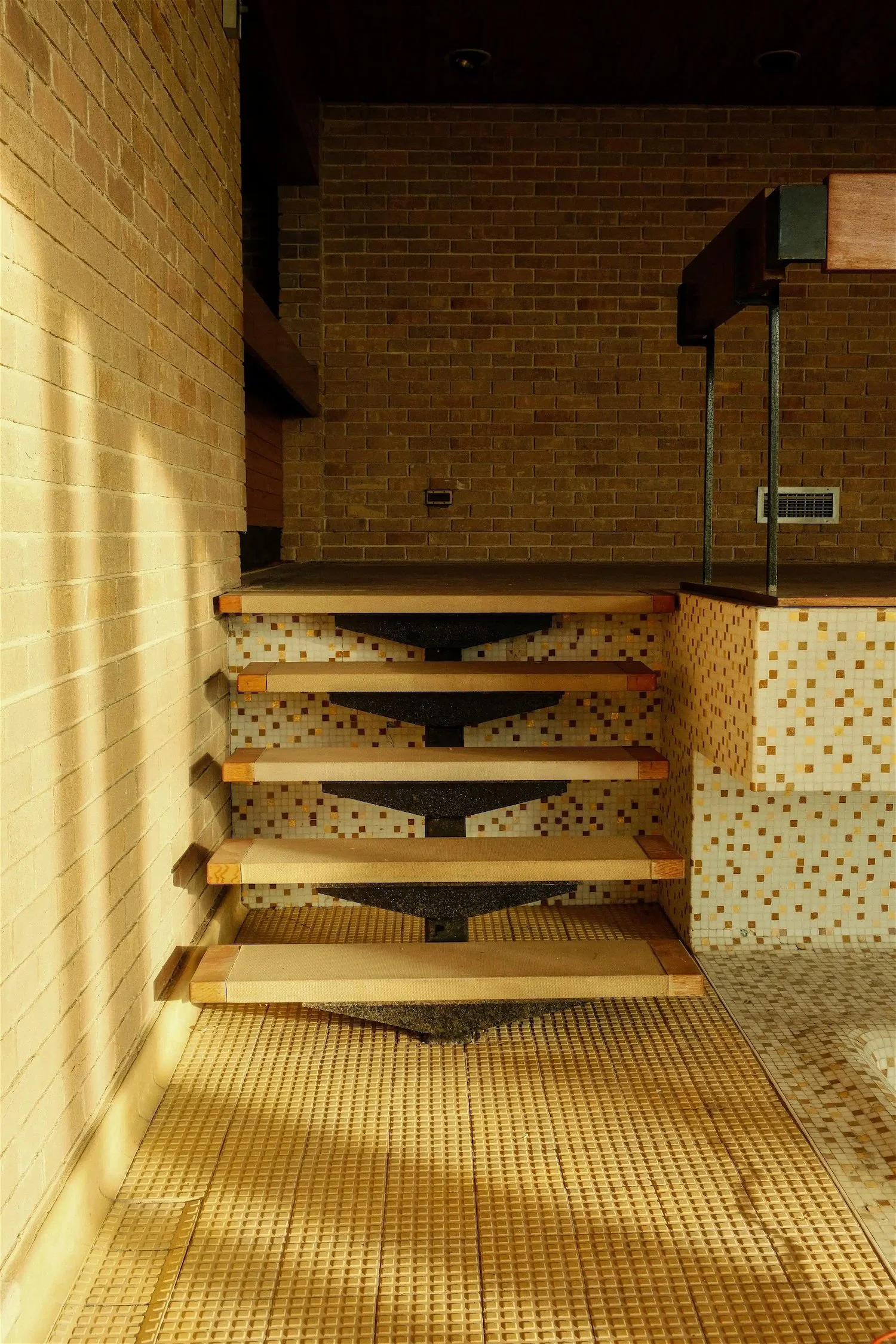 Interior of a building showing a staircase with wooden steps and black supports, with brick and mosaic tile walls and a tiled floor.