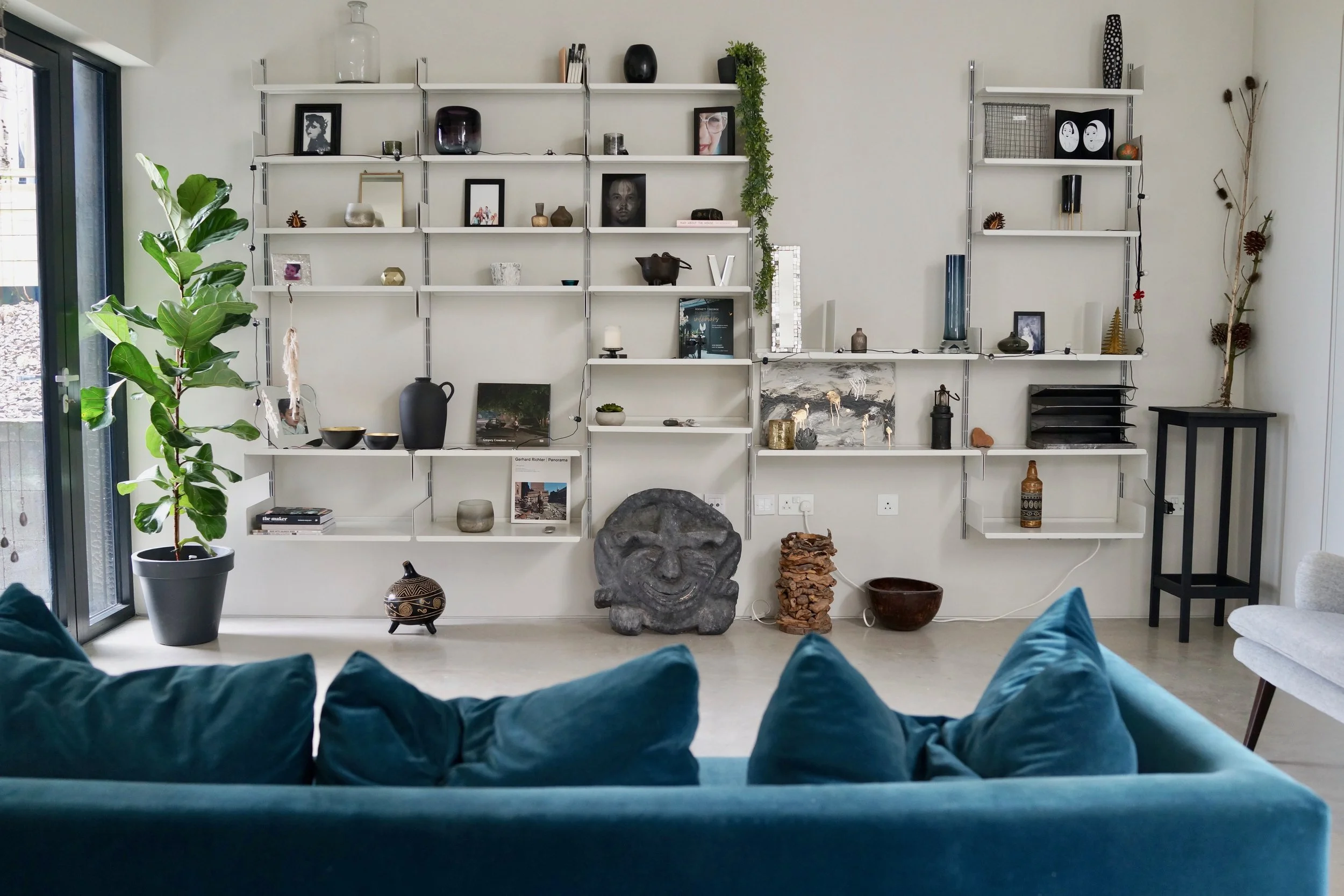 Living room with a white wall and modern white shelving decorated with various art pieces, vases, and books. A large green potted plant is on the left near a sliding glass door, with a blue sofa and teal cushions in the foreground. Decorative items o