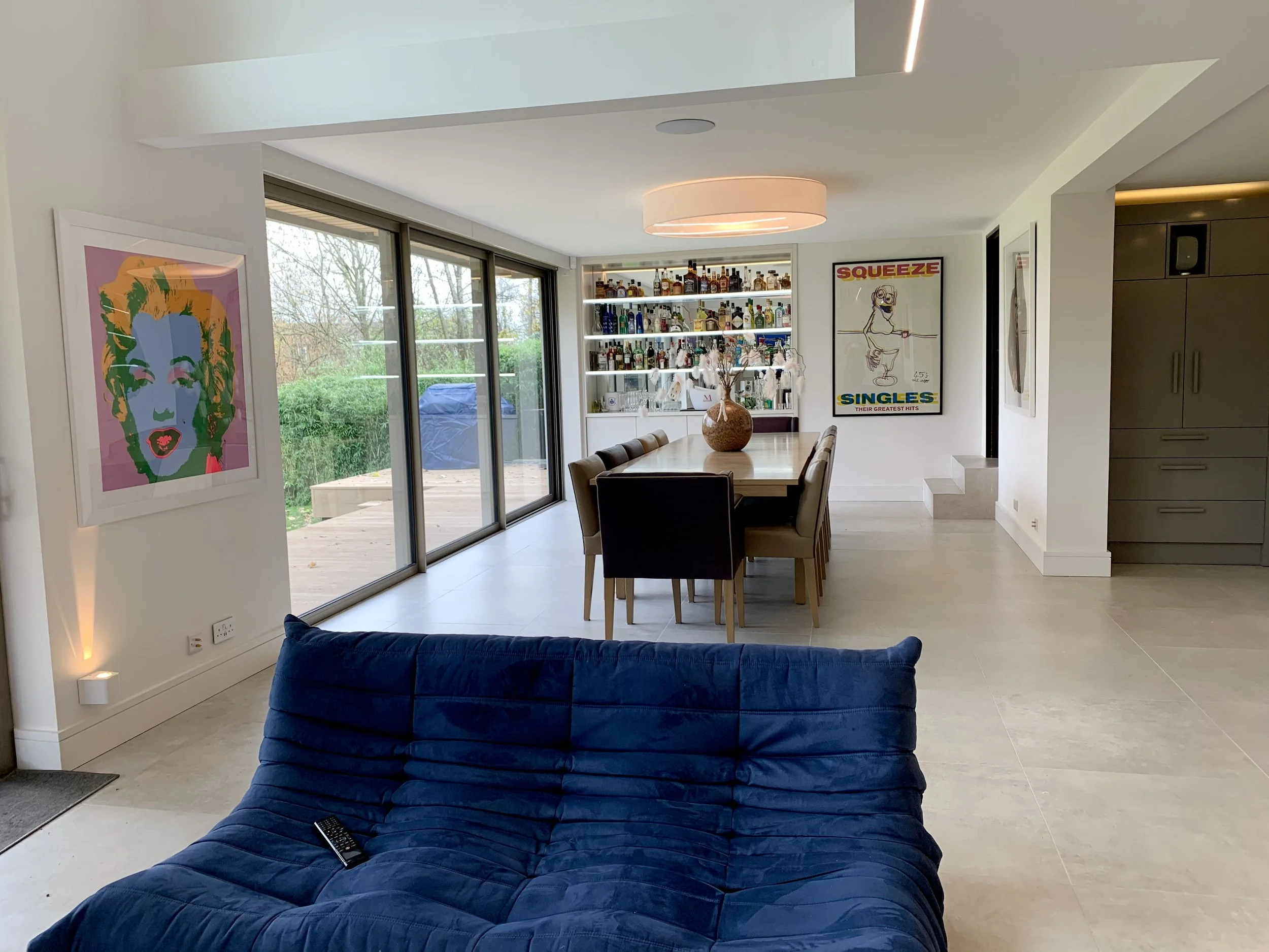 Modern dining room with large sliding glass doors, a wooden dining table with eight chairs, a bar with bottles of alcohol, and colorful art on the walls, including a portrait of Marilyn Monroe and a Squeeze concert poster.