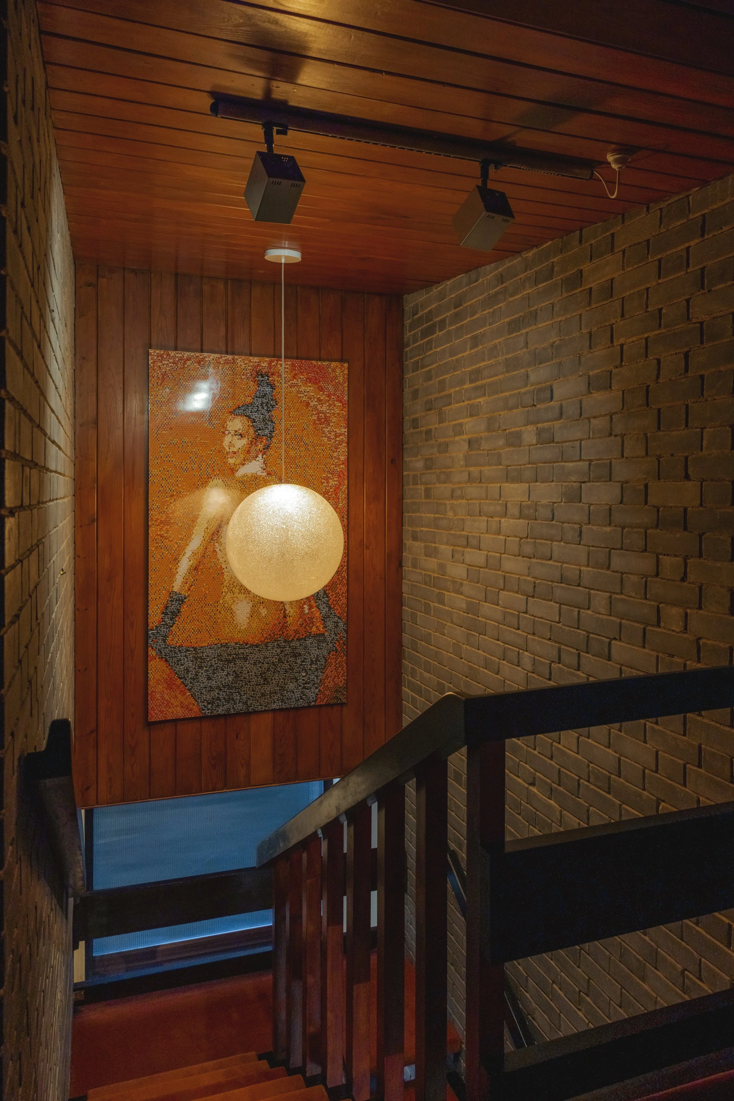 Wood-paneled ceiling with two mounted speakers and a hanging round pendant light illuminating a mosaic artwork of a woman with dark hair on a wooden wall, next to a brick wall, descending staircase with a wooden handrail.