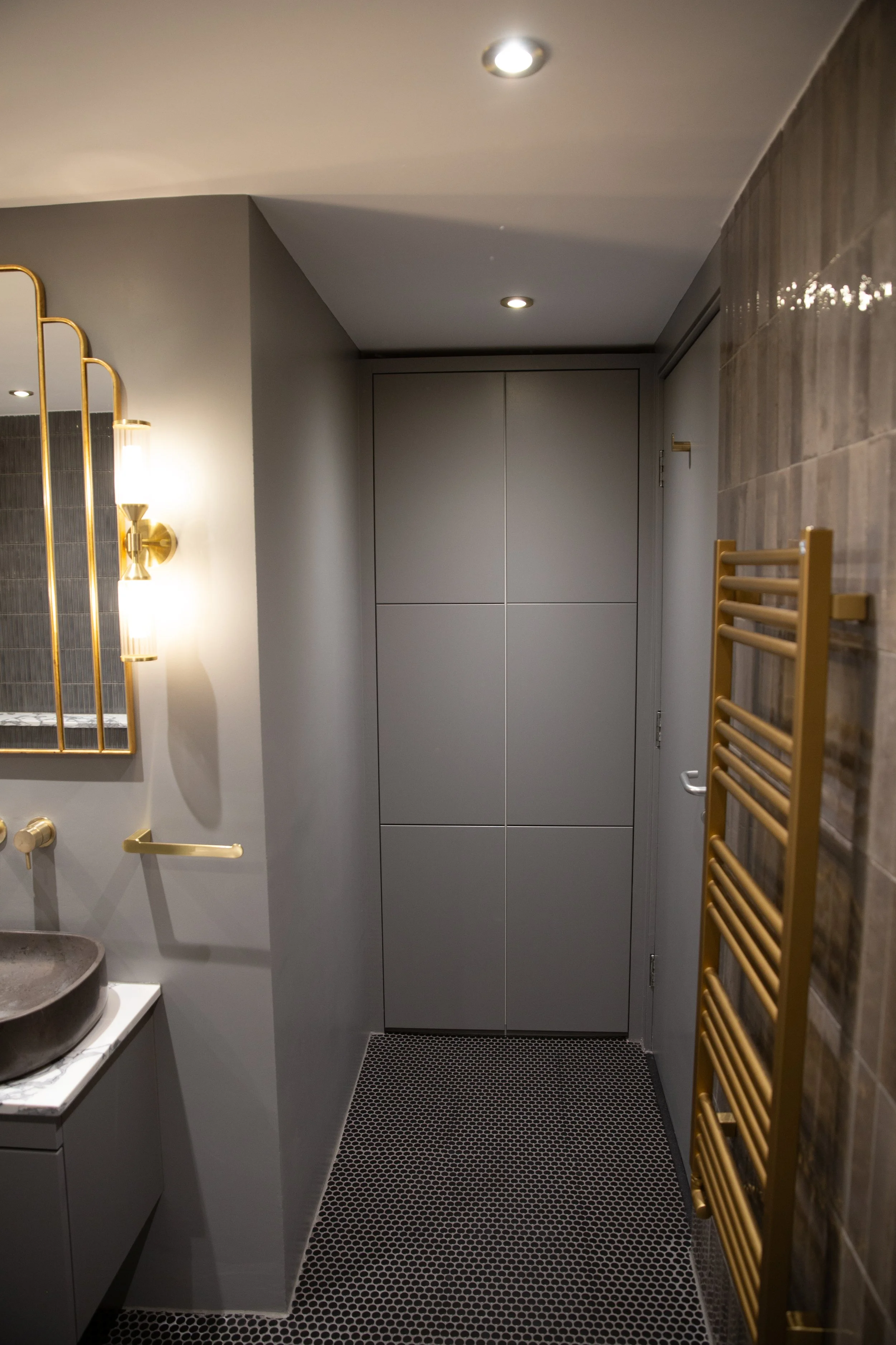 A modern bathroom corner with gray cabinets, a gold towel rack, a mesh black and white floor, and a light gray wall with a mirror and a gold light fixture.