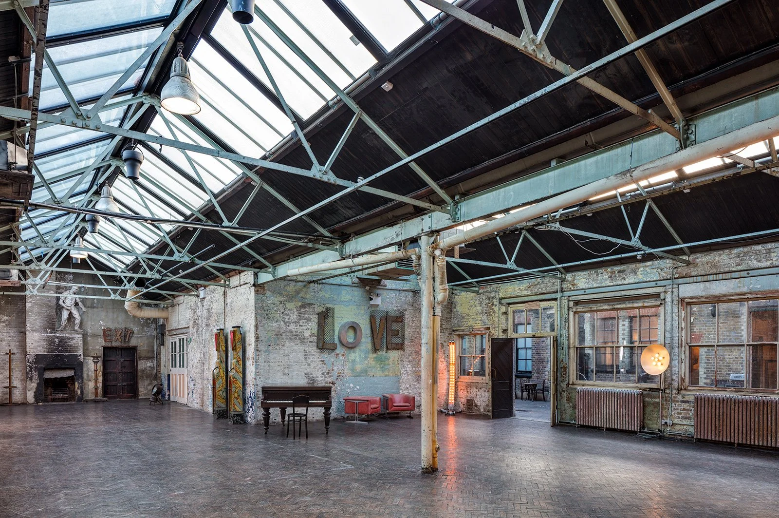 An industrial loft with exposed brick walls, large windows, and a glass roof. Inside, there are a piana, red armchairs, and a wall with the word LOVE spelled out with metallic letters. There is also a sculpture and some colorful artwork.