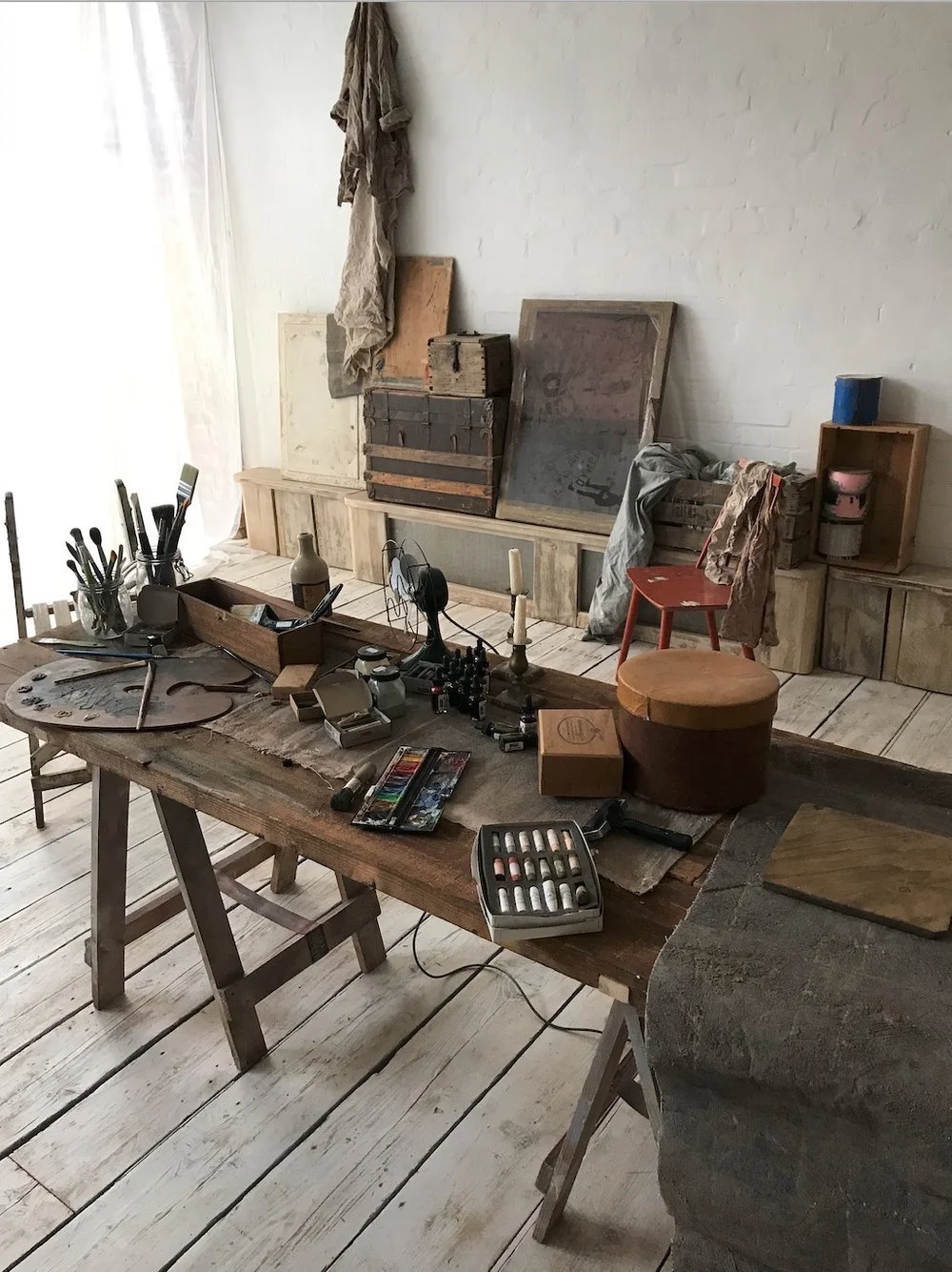 A rustic art studio workspace with a wooden table holding art supplies, including paints, brushes, and a palette. In the background, there are art frames, wooden boxes, and fabric on a raised platform against a white brick wall and a sheer curtain al