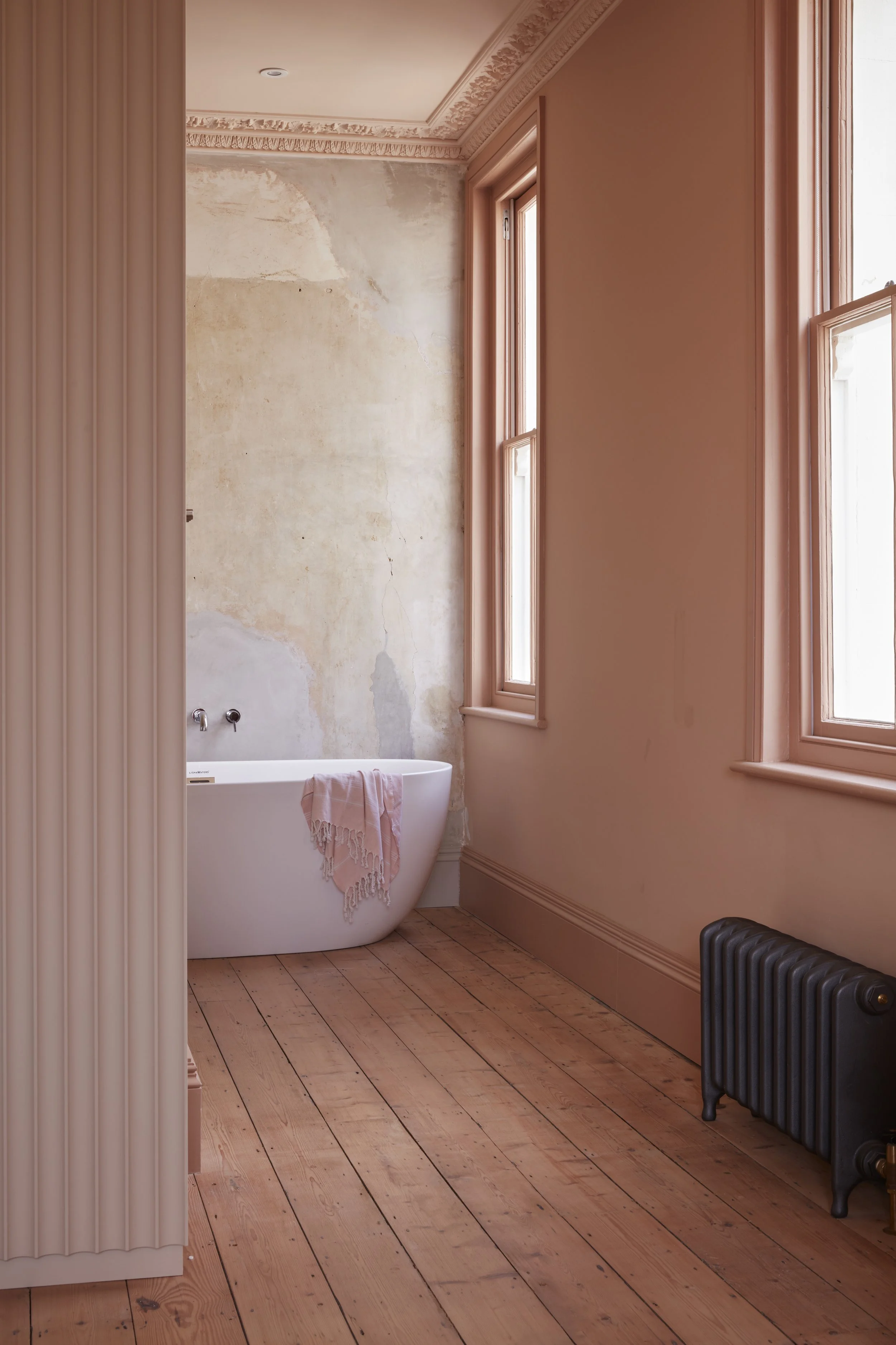 A bathroom with pink walls and wooden floors. There is a clawfoot bathtub with a pink towel draped over it near weathered wall with peeling paint. The room has two large windows with wooden frames and a black cast iron radiator.