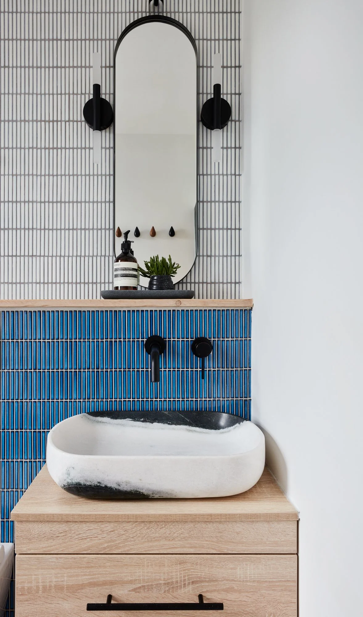 Modern bathroom vanity with a stone sink, wall-mounted black faucet and knobs, a mirror with black wall lamps, and decorative items including a plant and soap bottle against a background of colorful tiled walls.