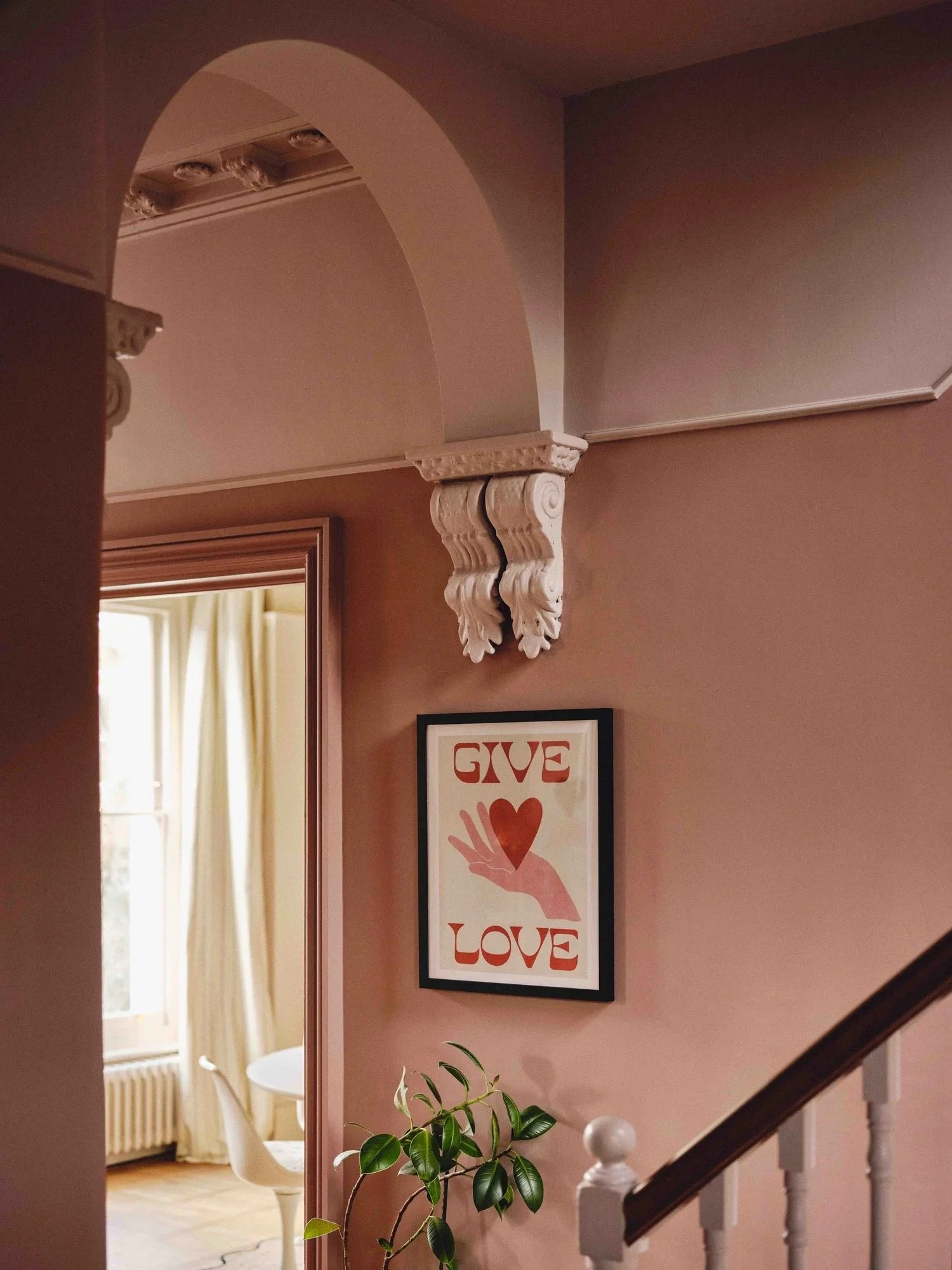 Decorative interior corner of a house with an arch, a framed print with the words 'GIVE LOVE' and a heart, a plant, and a staircase.