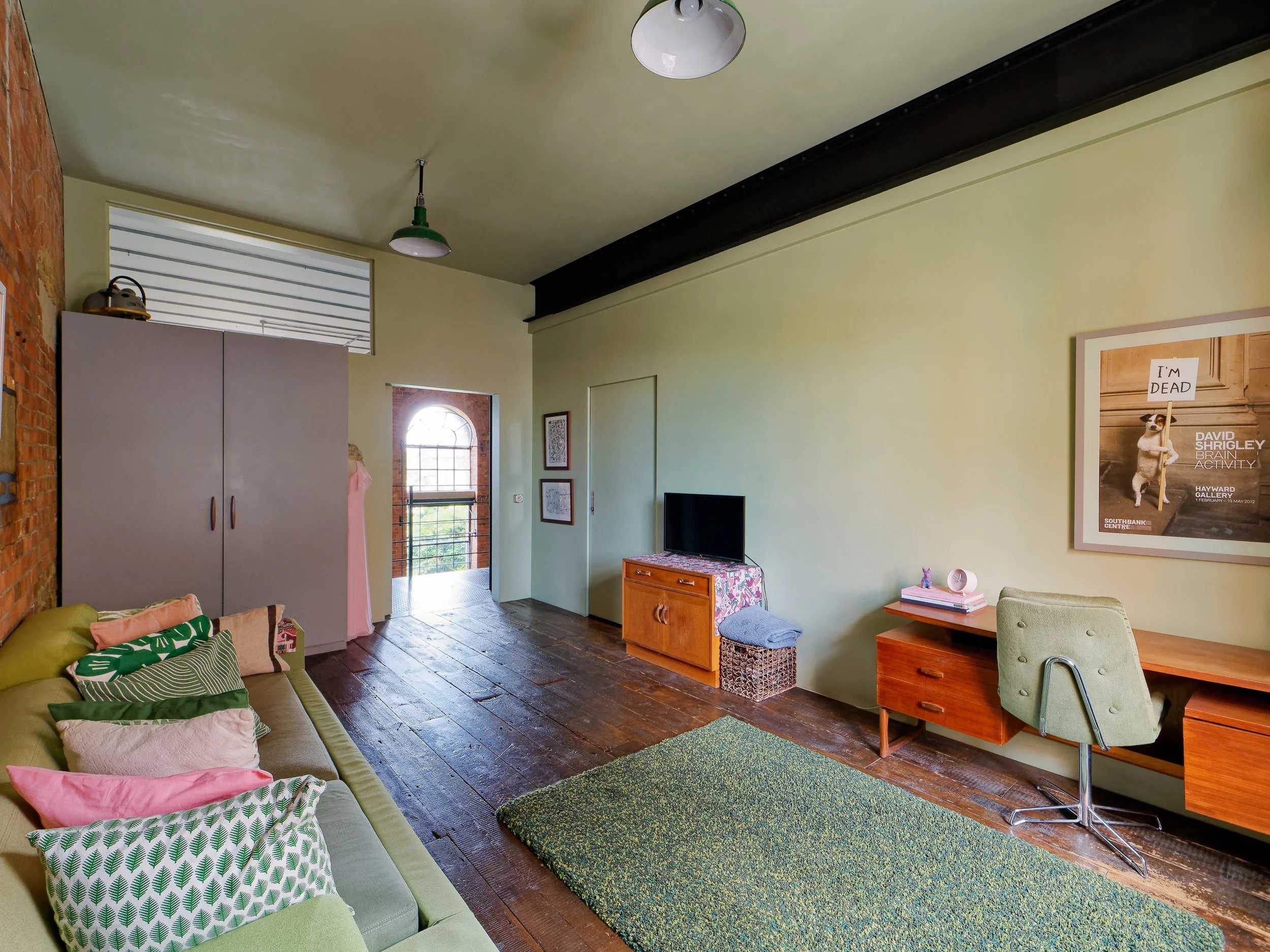 Living room with green upholstery sofa with multicolored pillows, a wooden desk with a green office chair, a flat-screen TV on a small wooden cabinet, framed art on the pale yellow wall, and a large window with a brick arch.