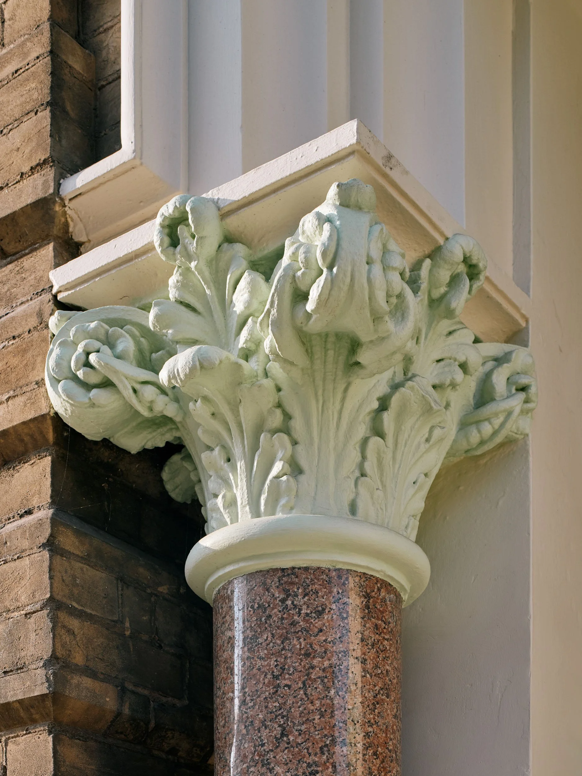 Decorative architectural column capital with ornate carved foliage, mounted on a granite cylindrical shaft, belonging to a building's exterior.