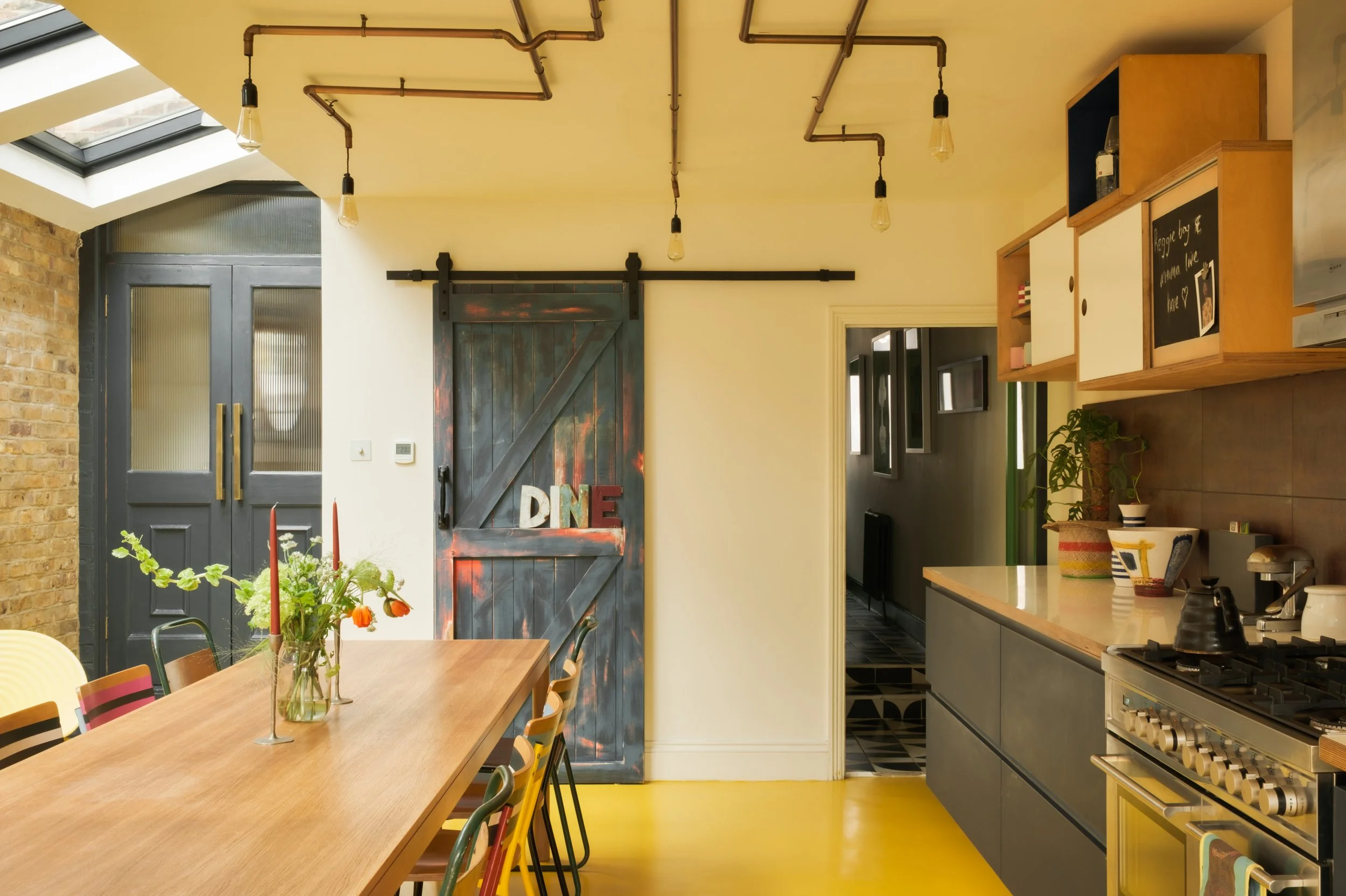 Jeanette kitchen with a long wooden dining table, a rustic sliding barn door with 'DINE' sign, exposed brick wall, yellow floor, and open shelving with decorative items.