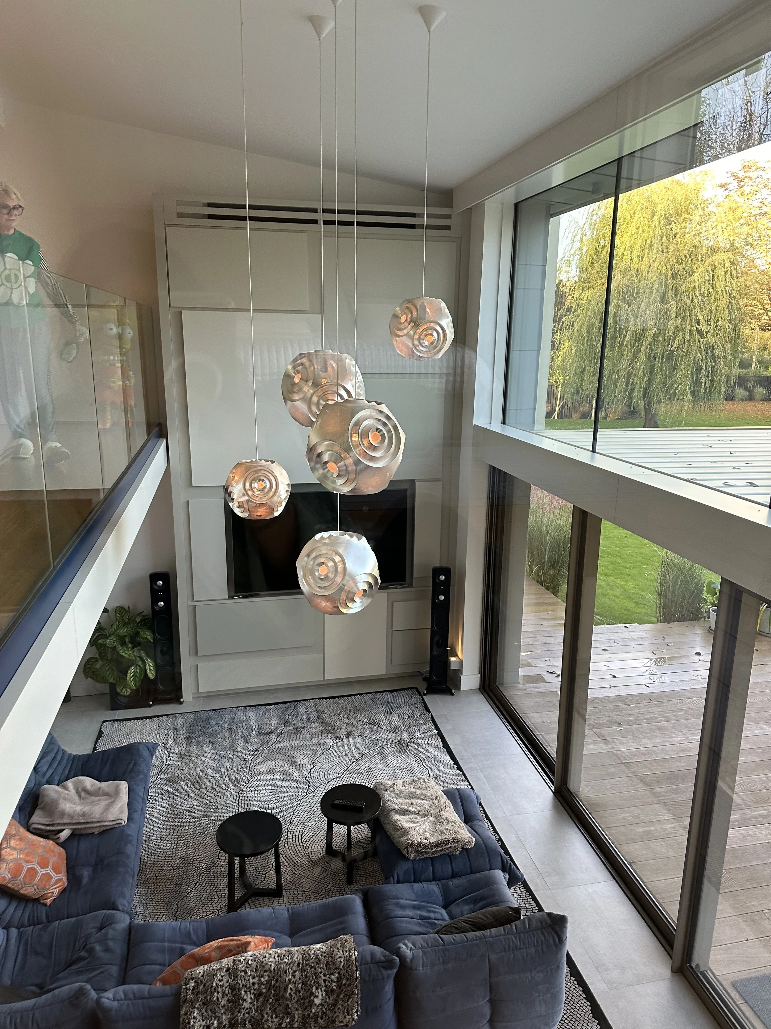Interior view of a modern living room with a high ceiling, featuring a white entertainment center housing a large flat-screen TV and tall black speakers on either side. A unique chandelier with six hanging metallic spiral-shaped lights is centered ab
